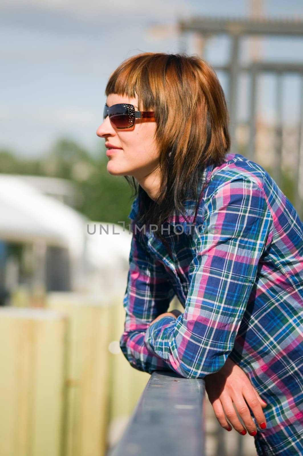 Side upper body view of a young brunette woman with plaid shirt and sunglasses leaning on a railing and looking to the side