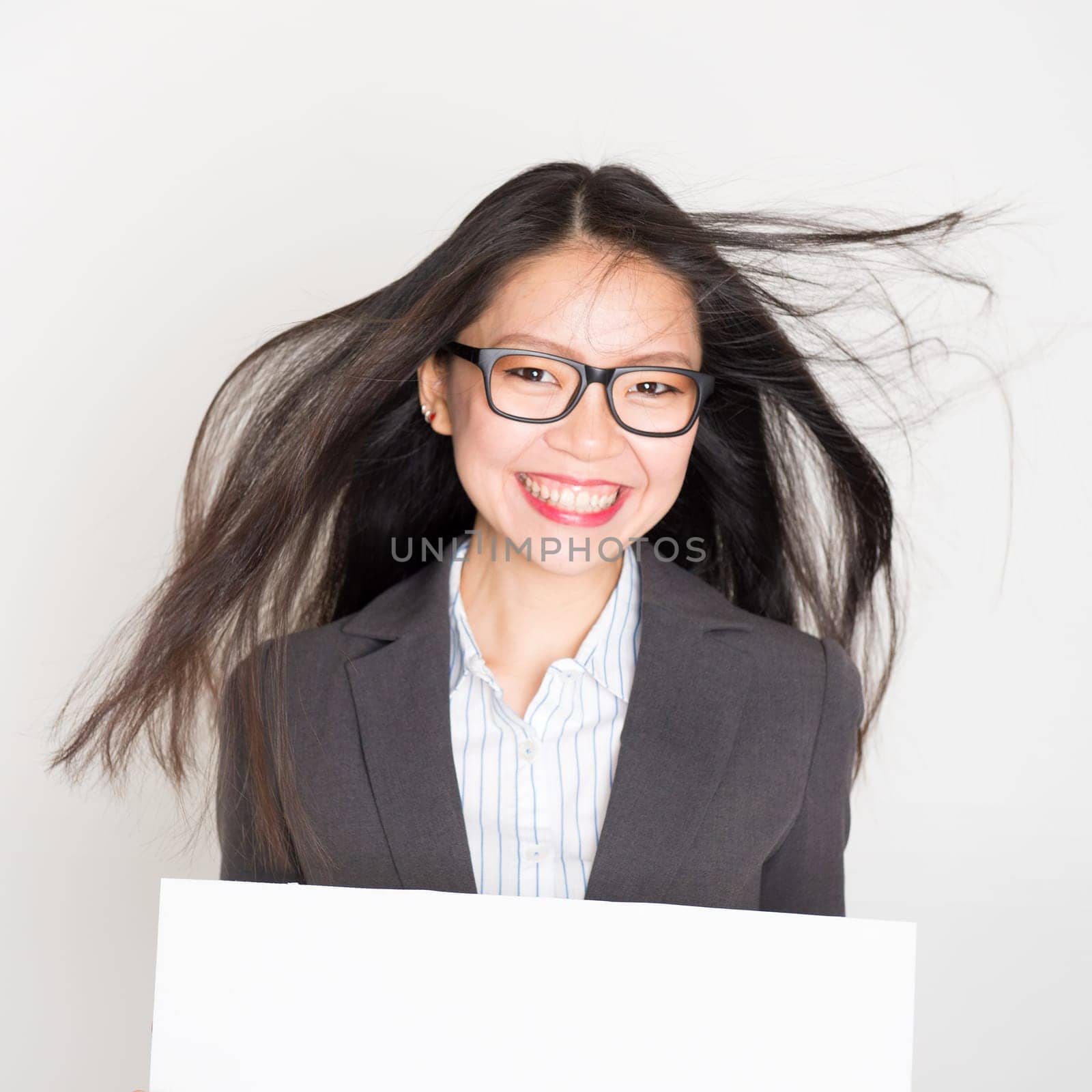 Portrait of Asian businesswoman in formalwear smiling and hand holding white blank paper card, standing on plain background.