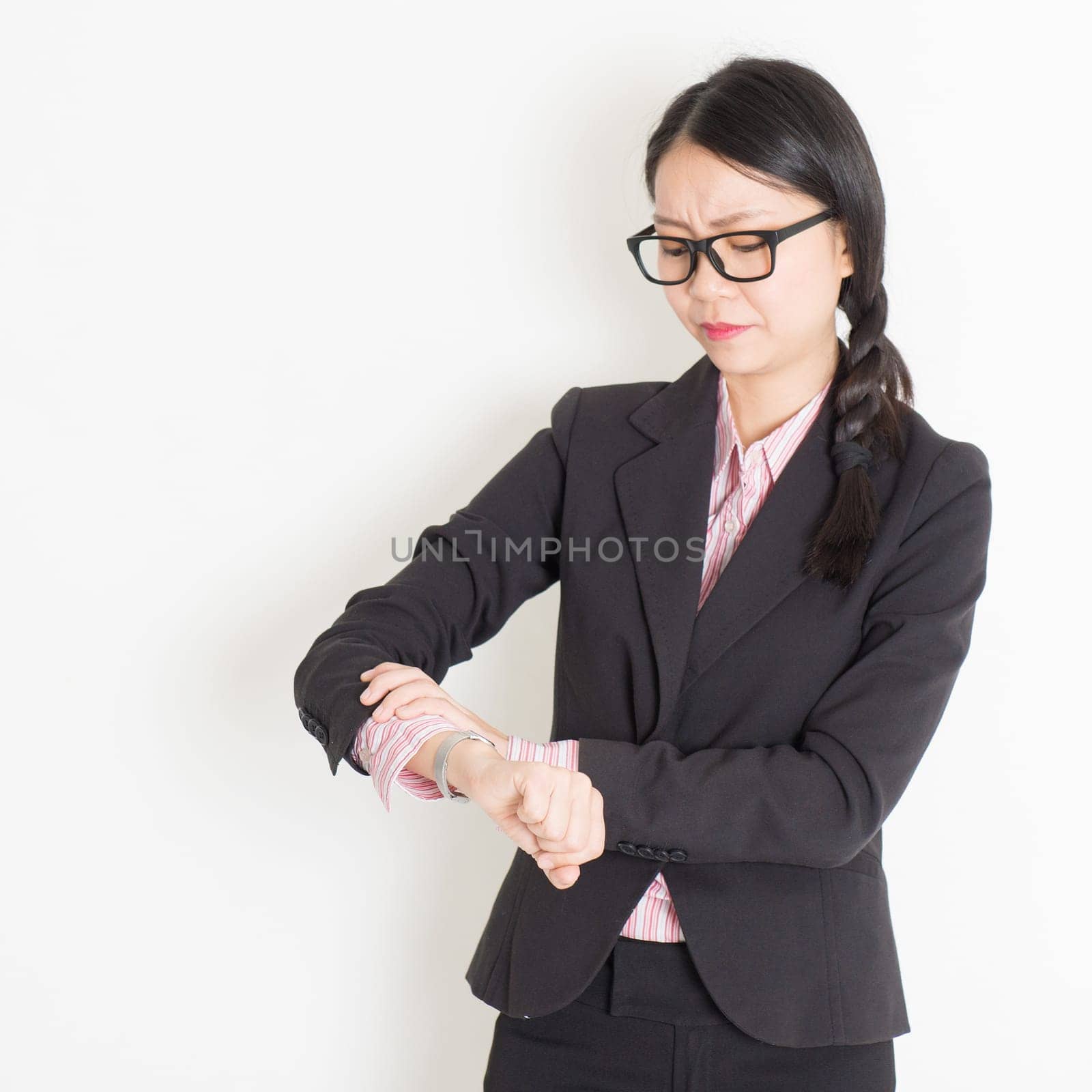 Young Asian businesswoman checks time on her wrist watch, late concept, standing on plain background.