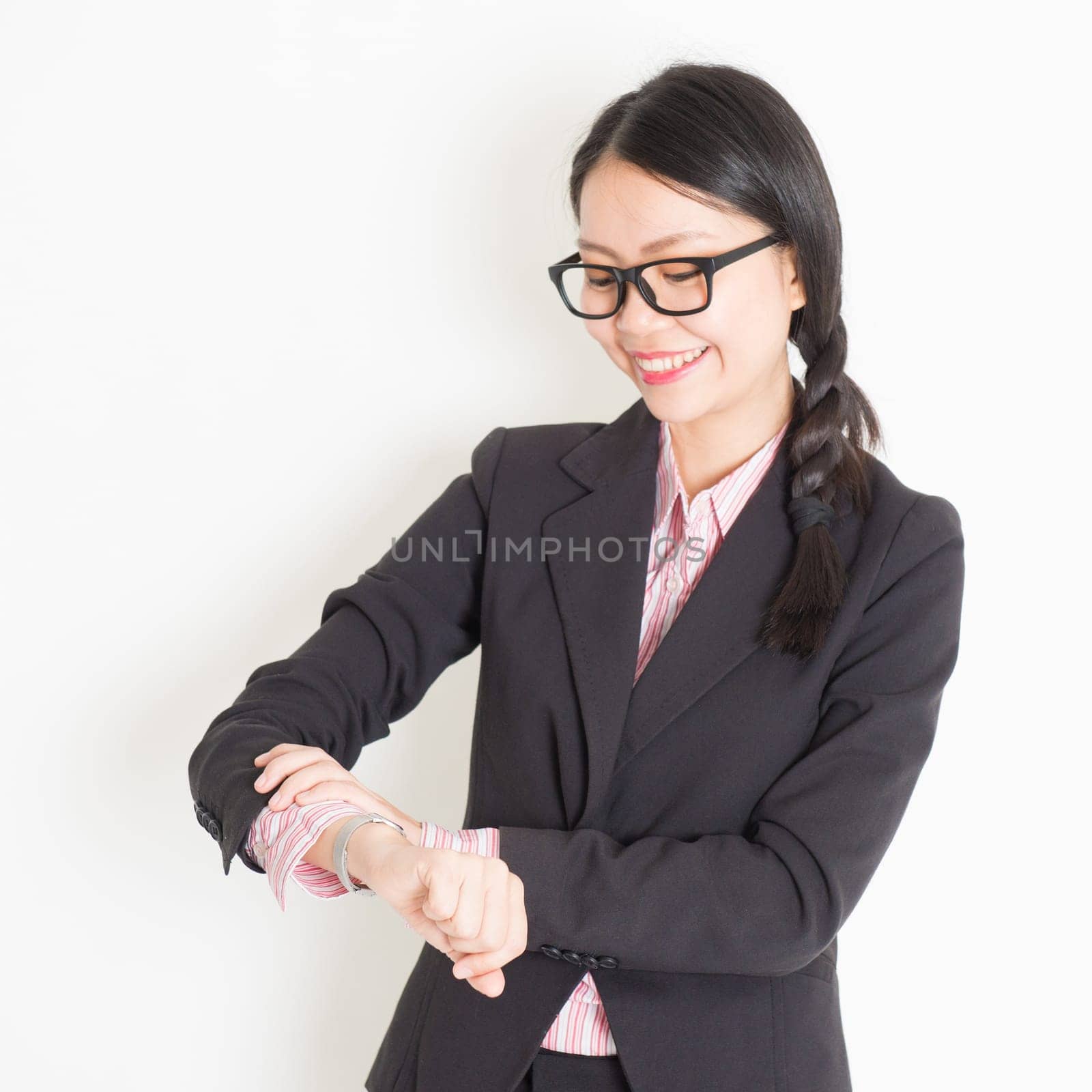 Young Asian businesswoman checks time on her wrist watch, standing on plain background.
