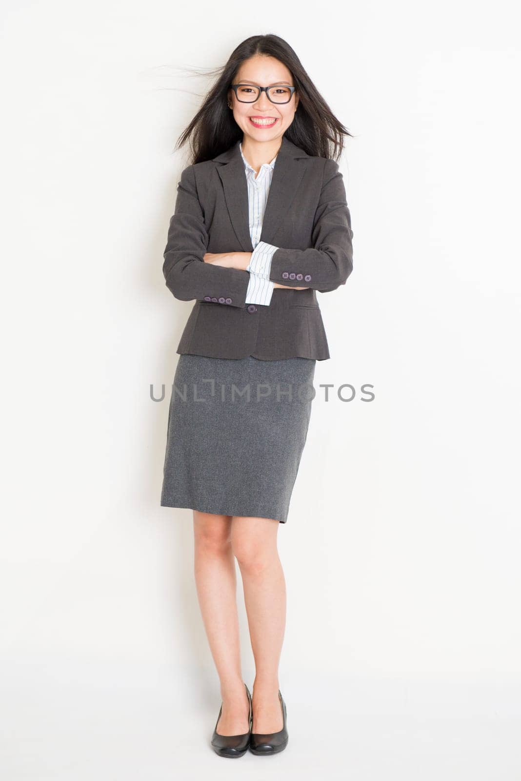 Portrait of Asian female business woman in formalwear smiling, full body standing on plain background.