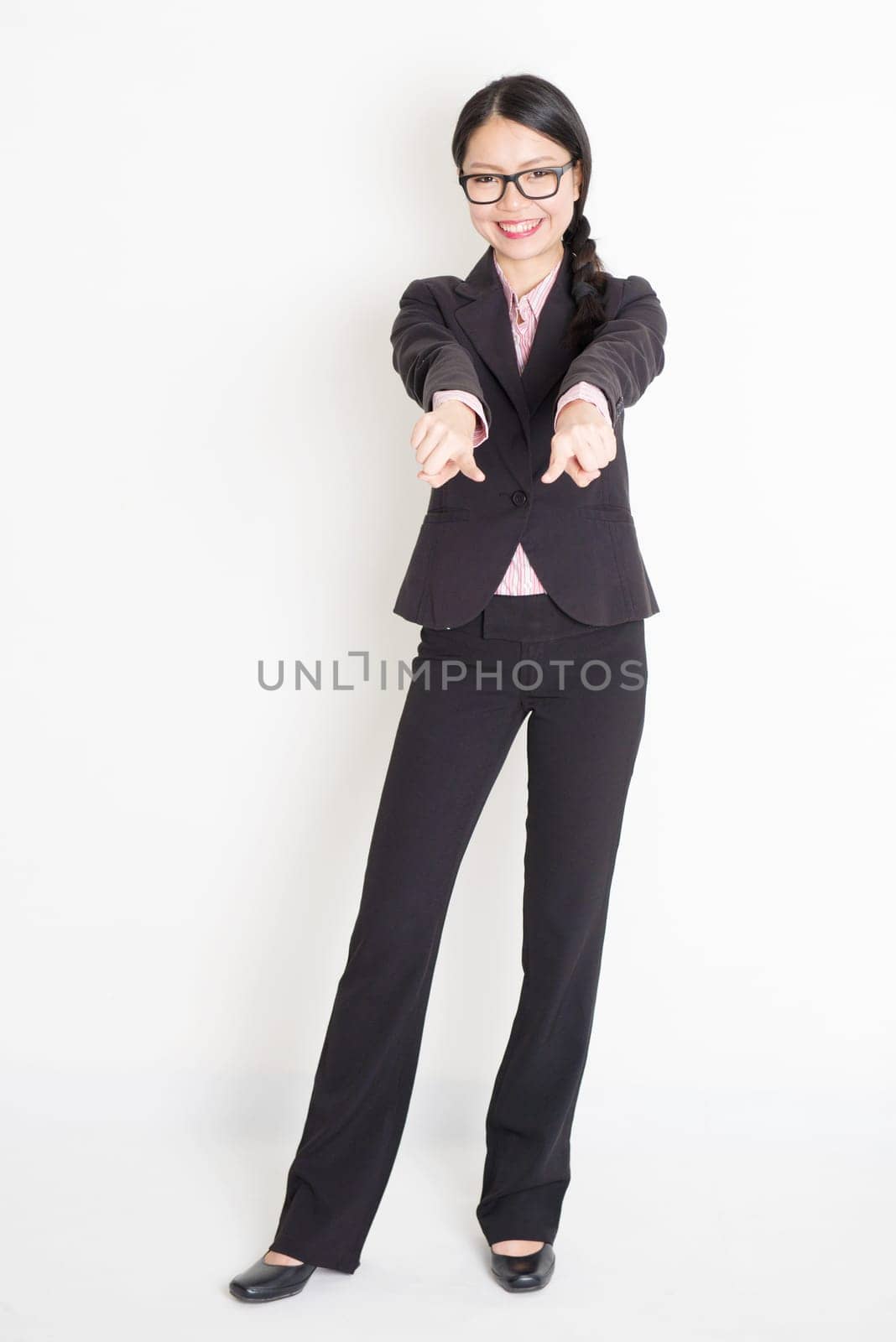 Full body portrait of young Asian businesswoman in formalwear fingers pointing on you and smiling, standing on plain background.