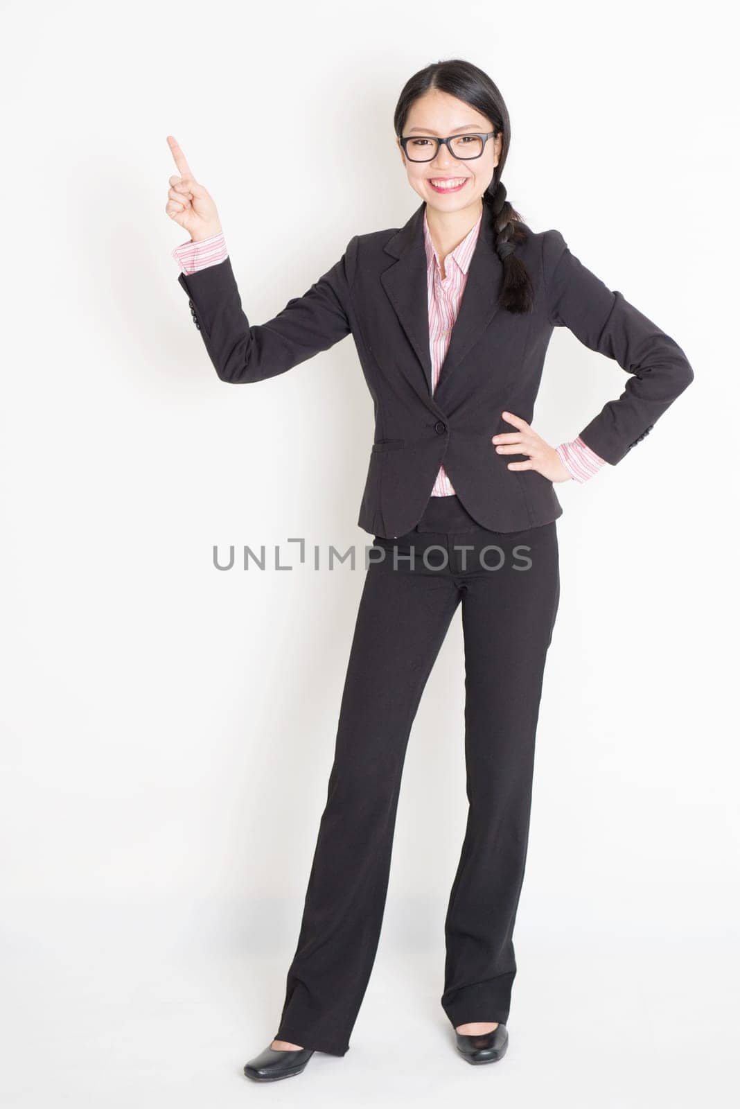 Full body portrait of young Asian businesswoman in formalwear fingers pointing at copy space and smiling, standing on plain background.
