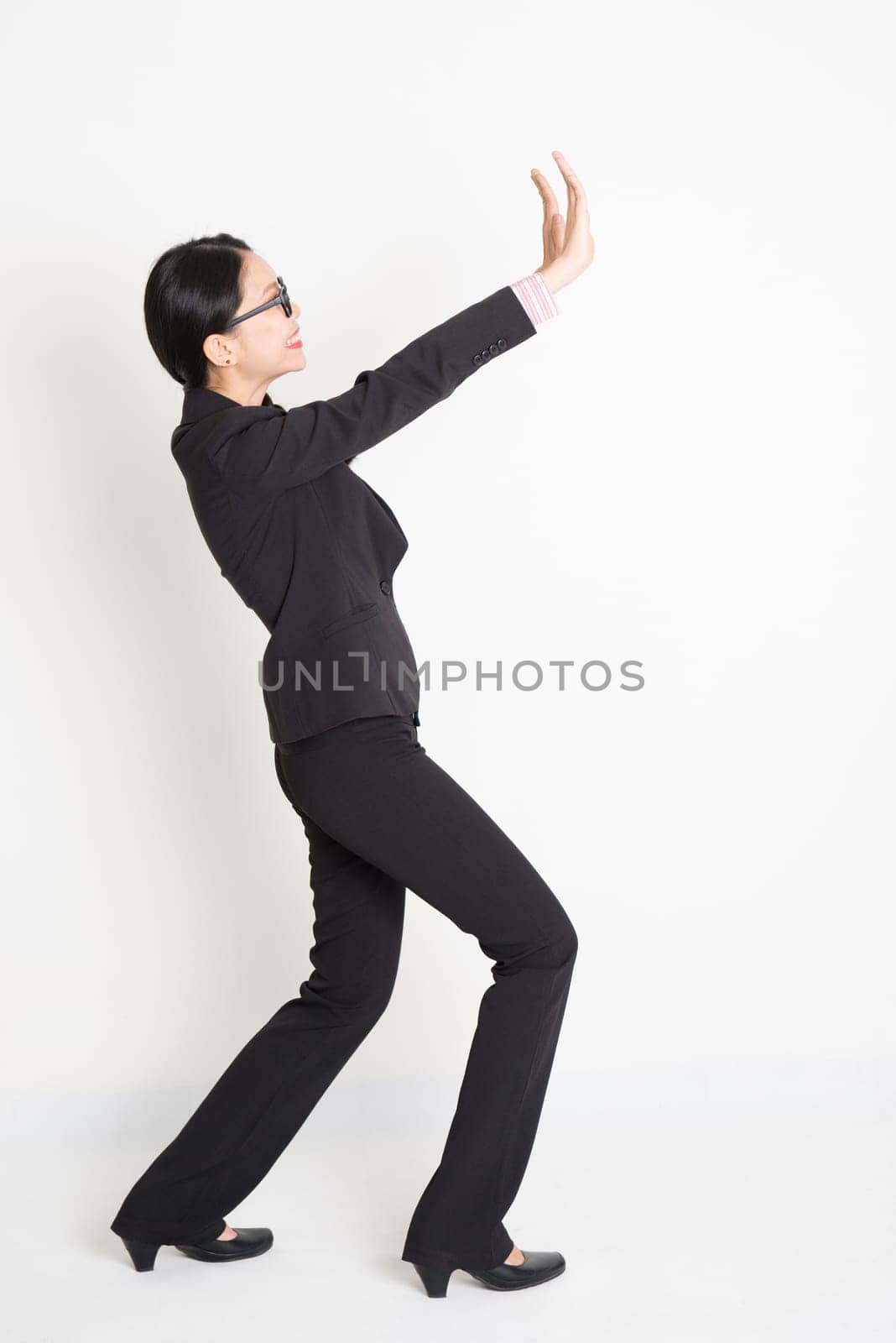 Full body portrait of young Asian businesswoman in formalwear hands pushing on blank space and smiling, standing on plain background.