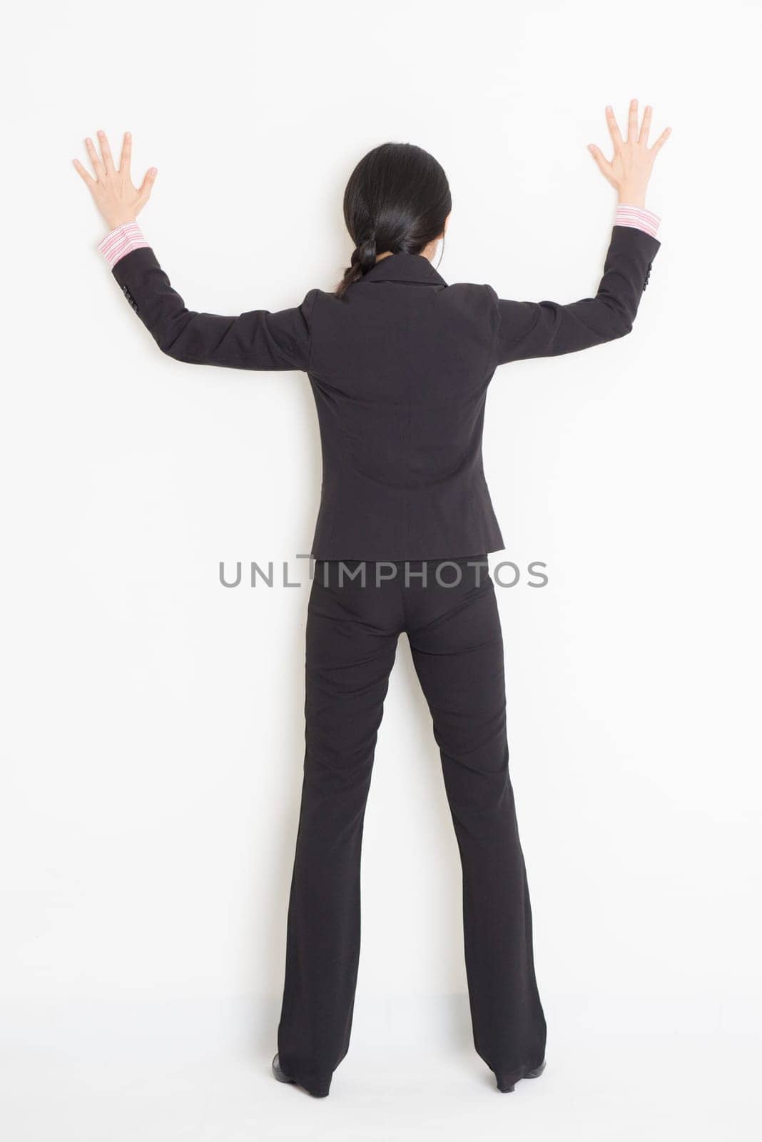 Full body back view of young Asian businesswoman in formalwear banging on wall, standing on plain background.