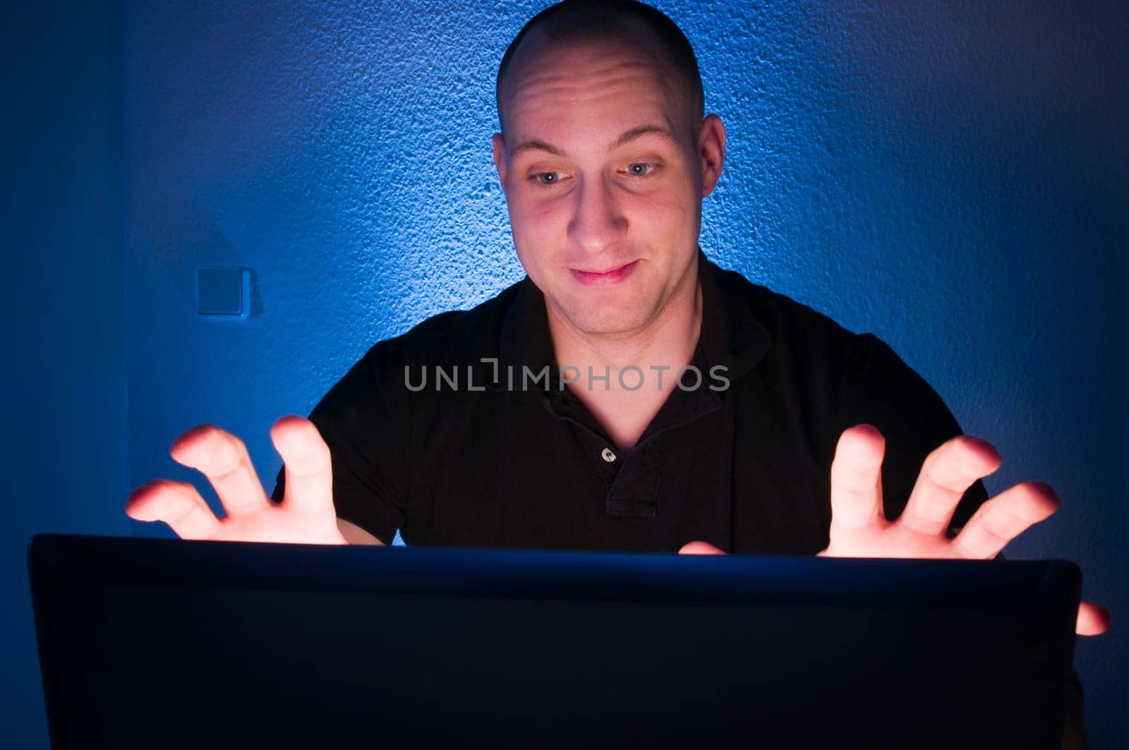 Head-and-shoulder shot of a young man sitting on a laptop monitor against a nightly blue background with a shocked expression on his face and frightened raised hands towards the monitor Camera position is behind the monitor towards the man's face
