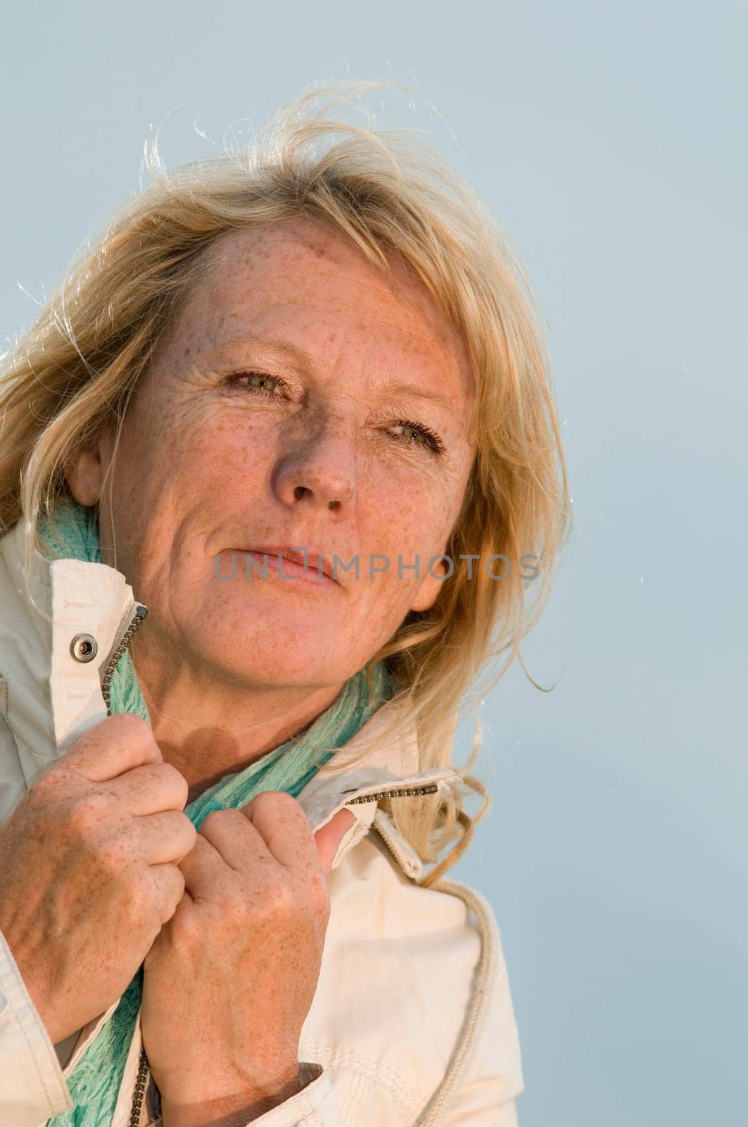 Upper body portrait of a blonde mature woman in a light jacket against an overcast sky looking dreamily to the side with her hands on the collar  Increased view