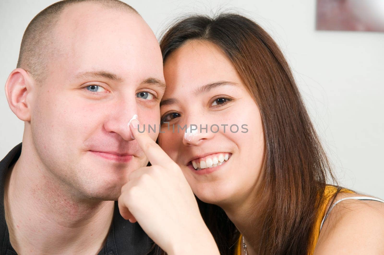 Head-and-shoulder portrait of a young couple dabbing white cream on each other's noses against a white background