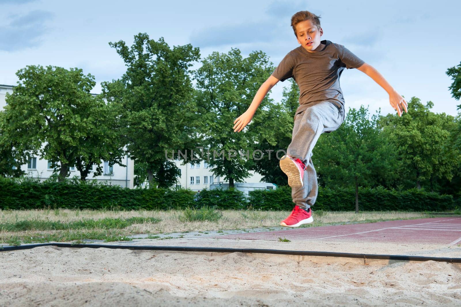 Side frontal full-body view of a male young teenager frozen while jumping into a long jump pit.