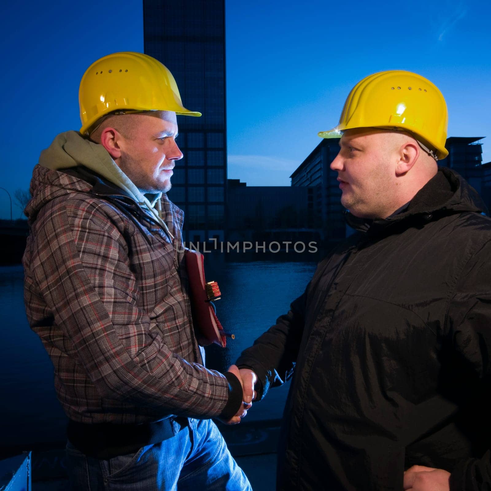 Upper body view in lower view of two men with yellow construction worker helmets reaching hands in front of a blue sky at night