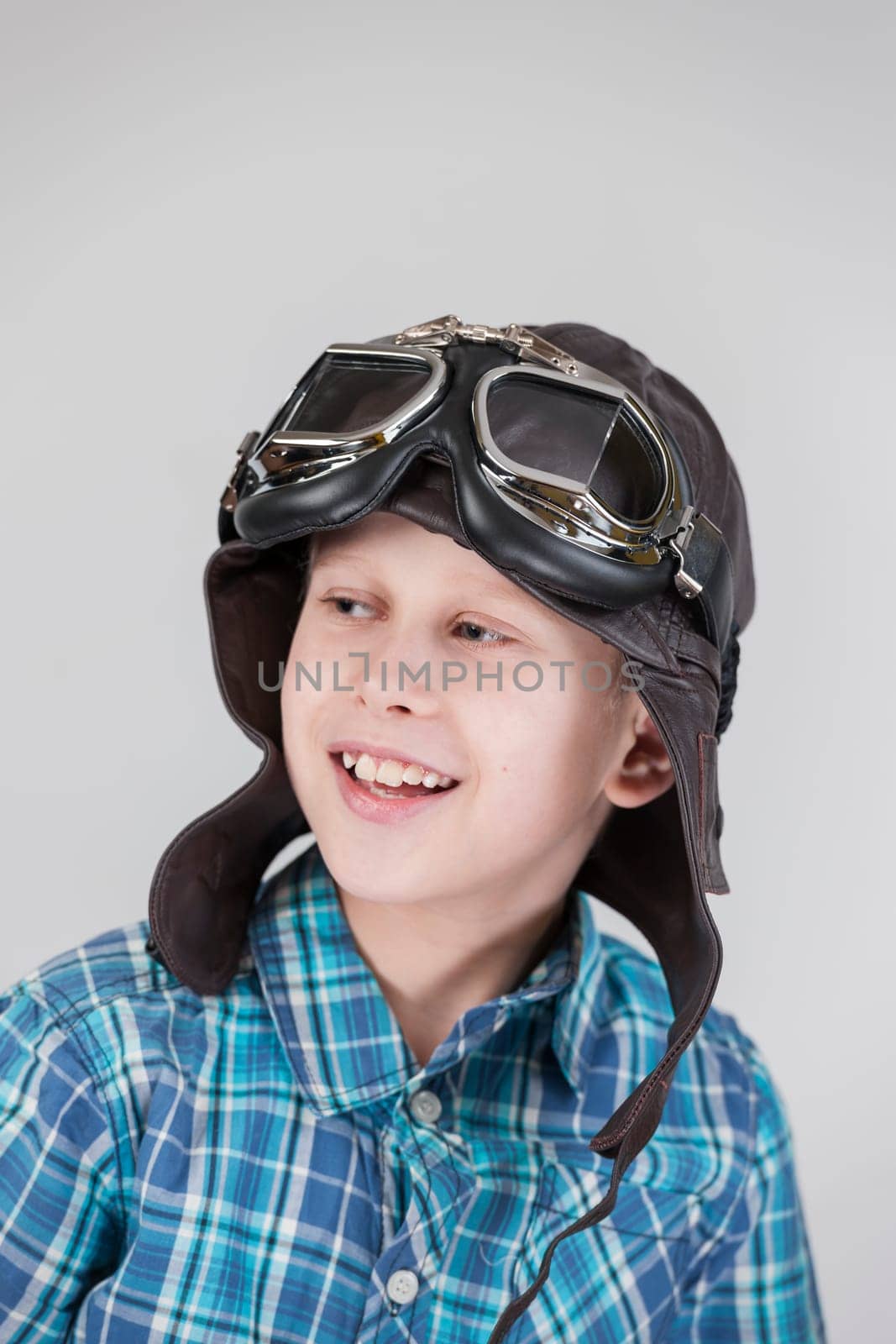 Against a grey background,the exposed upper body view of a ten-year-old boy in a blue-checkered short-sleeved shirt and brown leather cap with classic motorcycle glasses looks happily to the side.