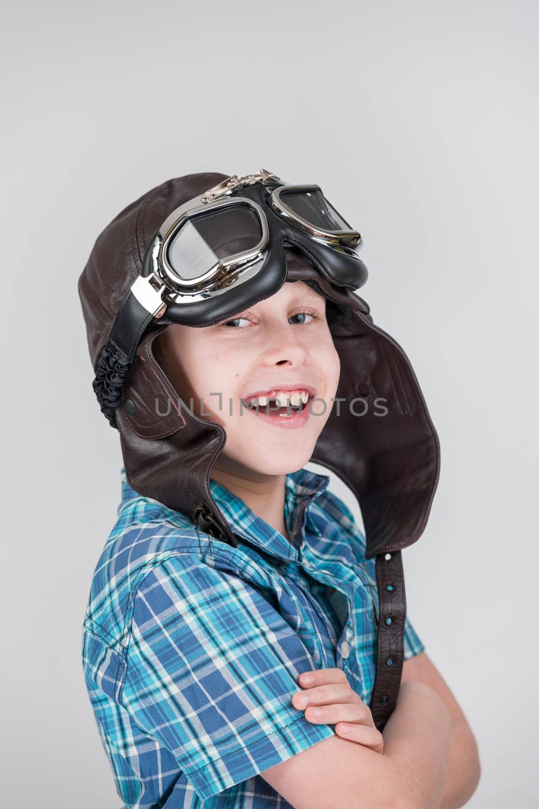 Against a grey background,the exposed upper body view of a ten-year-old boy in a blue-checkered short-sleeved shirt and brown leather cap with classic motorcycle glasses smiling joyfully into the camera.