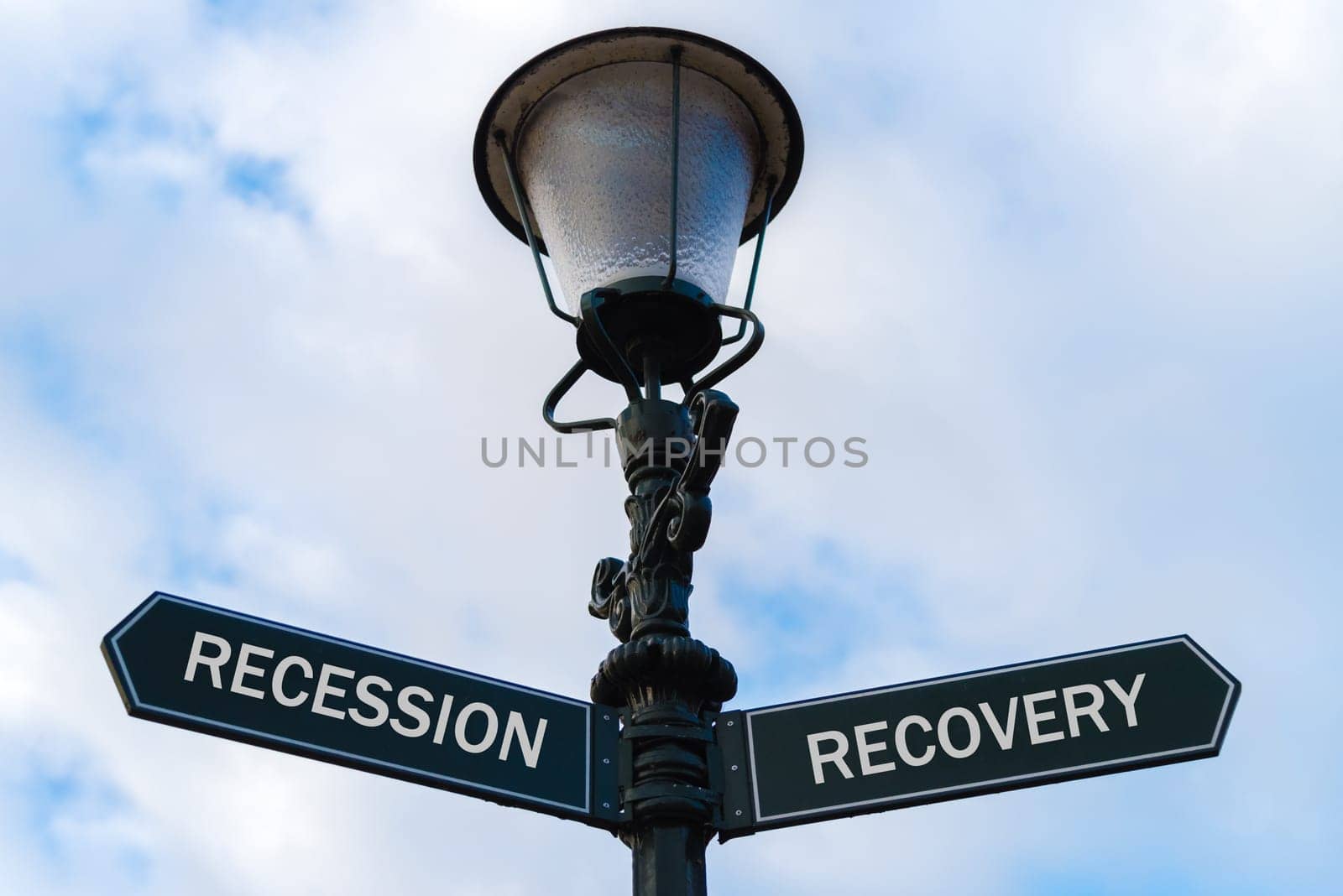 Street lighting pole with two opposite directional arrows over blue cloudy background. Recession versus Recovery concept.
