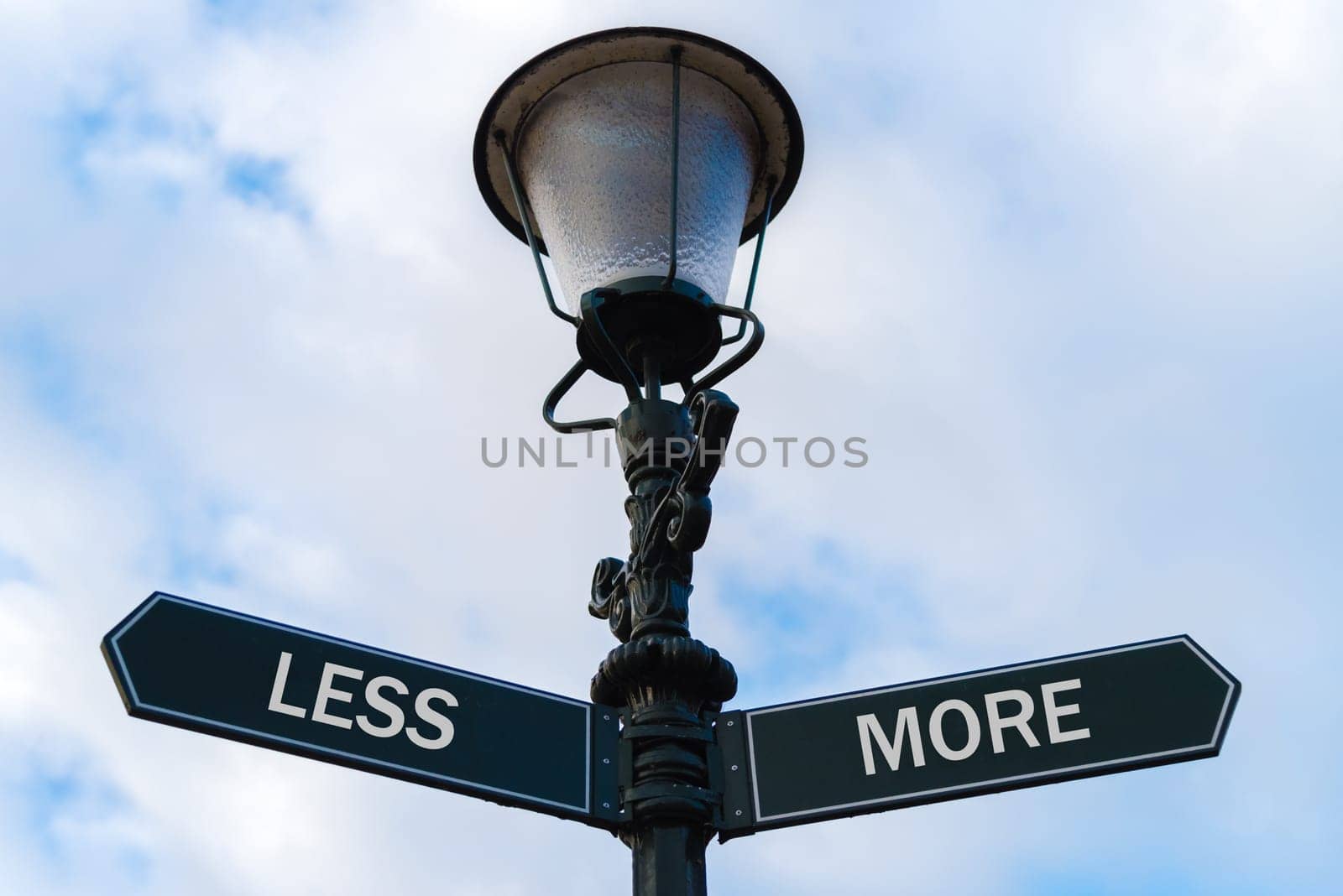 Street lighting pole with two opposite directional arrows over blue cloudy background. Less versus More concept.