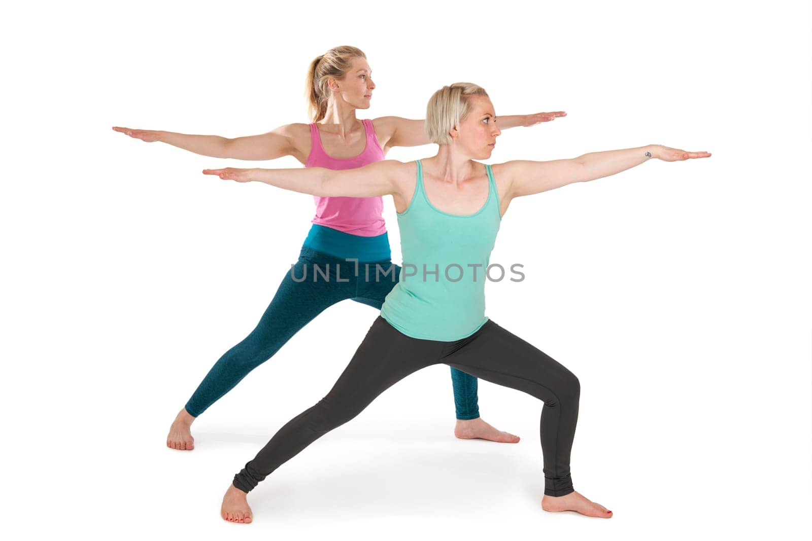 Full-body view of two young blonde women during parallel yogy exercise "the warrior"" against a white background with slight shadows"