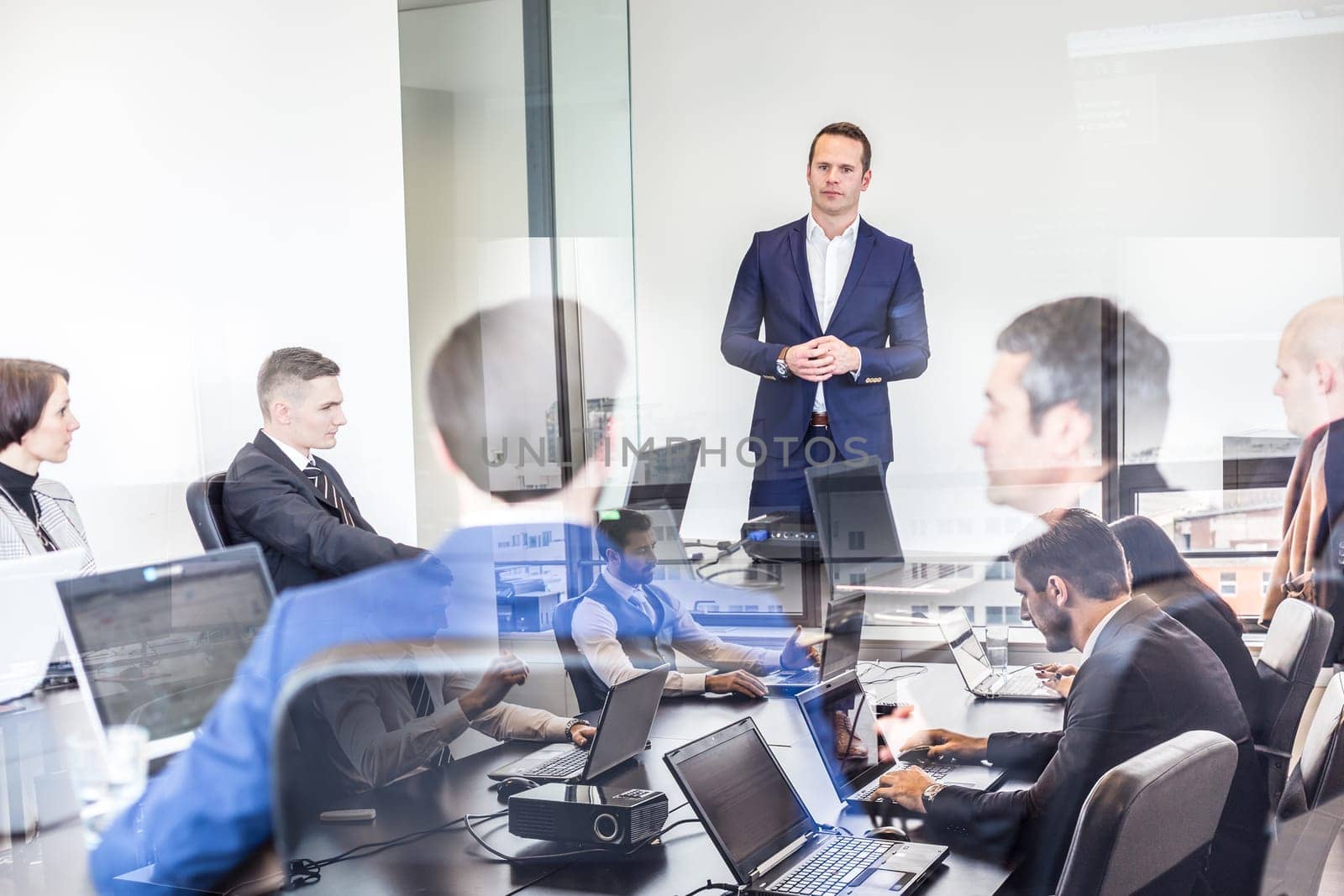 Successful team leader and business owner leading business meetingin corporate office. Business people working on laptops in foreground and glass reflections. Business and entrepreneurship concept.