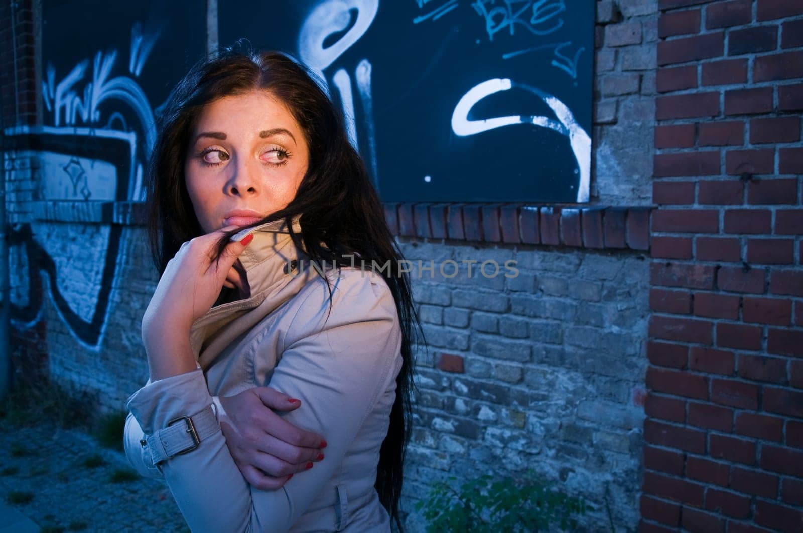 Young black-haired woman on a brick wall sprayed with graffiti in the evening illuminated with artificial light looking back anxiously.