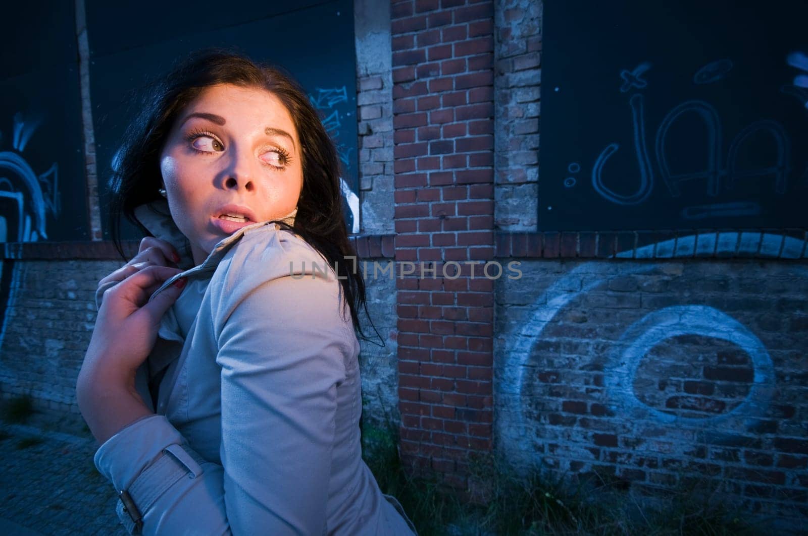 Young black-haired woman on a brick wall sprayed with graffiti in the evening illuminated with artificial light looking back anxiously.