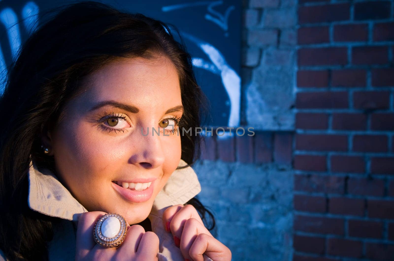 Young black-haired woman on a brick wall sprayed with graffiti illuminated with artificial light in the evening.
