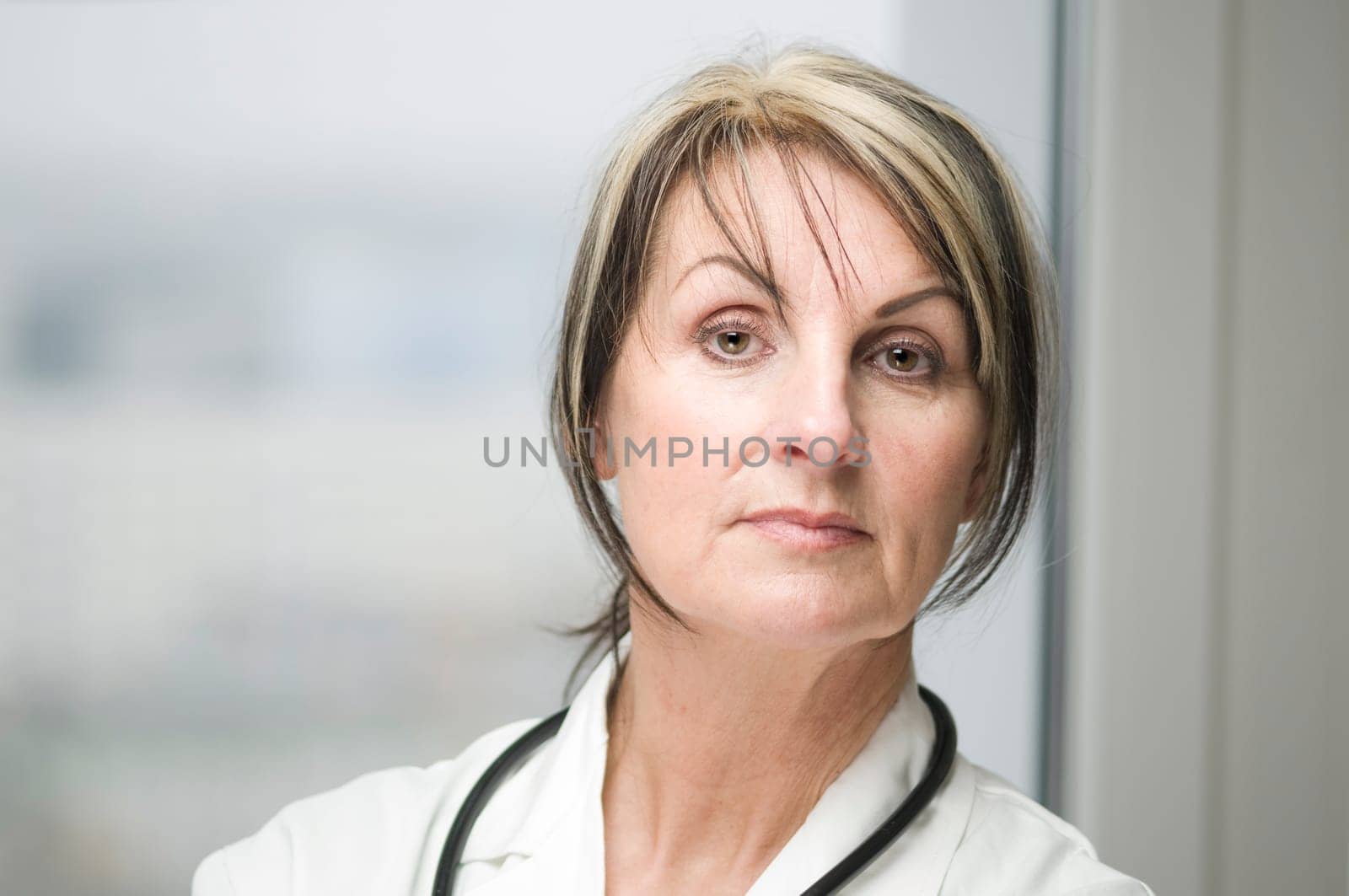 Frontal head-and-shoulder portrait of a middle-aged woman in a white coat with a stethoscope standing in front of a window and looking seriously into the camera.