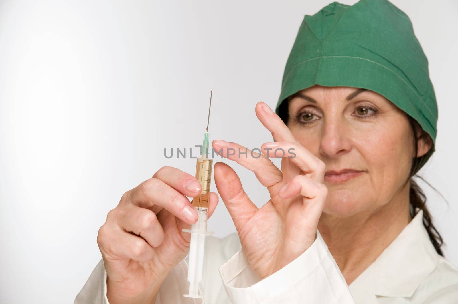 Head-and-shoulder view of a middle-aged woman with a green surgical hood against a white background holds a filled syringe in front of her.