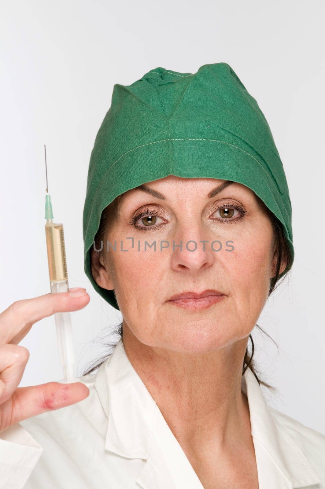 Head-and-shoulder view of a middle-aged woman with a green surgical hood against a white background holds a filled syringe in front of her.