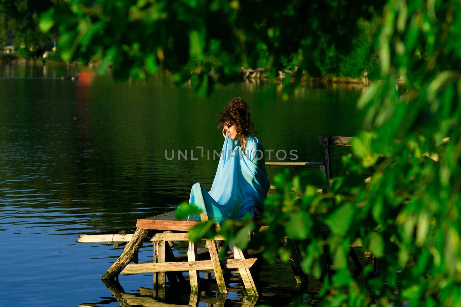 Extremely beautiful girl with perfect professional hairstyle.Nimf near water lying on the sun.Nymph,diva,goddess sits near the water,lake,river with blue dress,green trees,professional hairstyle. Summer greenery.Nimf near the water lying on the sun.