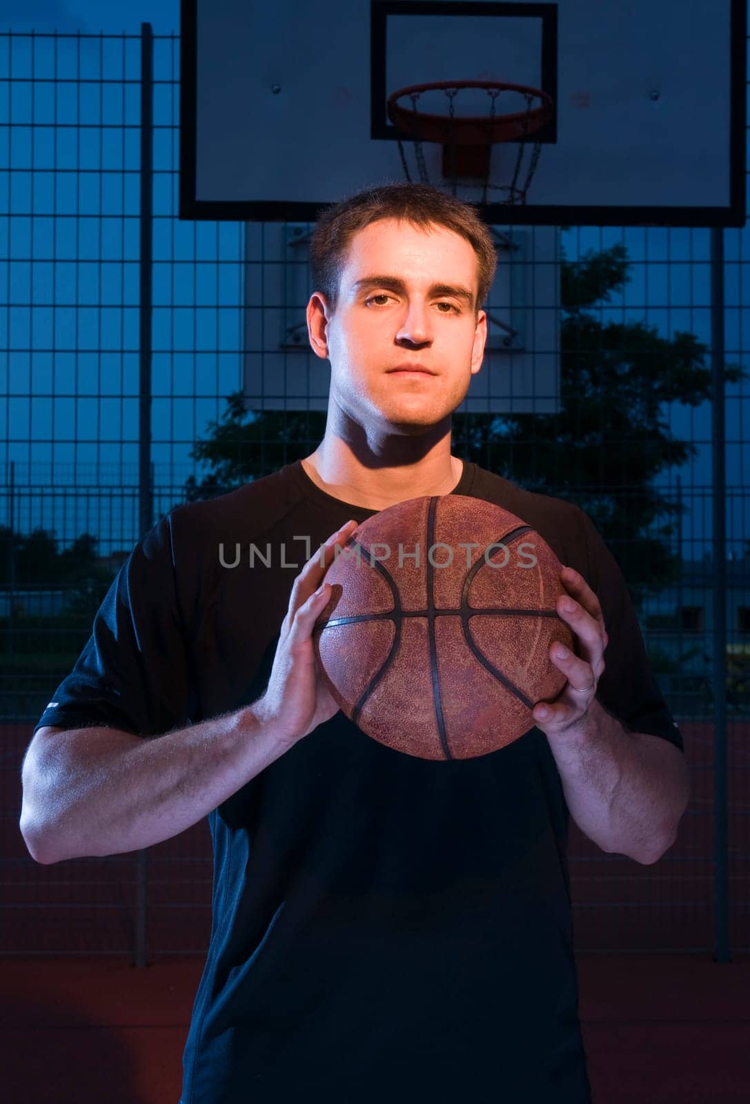 Frontal upper body view of a basketball player in the evening in the light with basketball held in front of him.