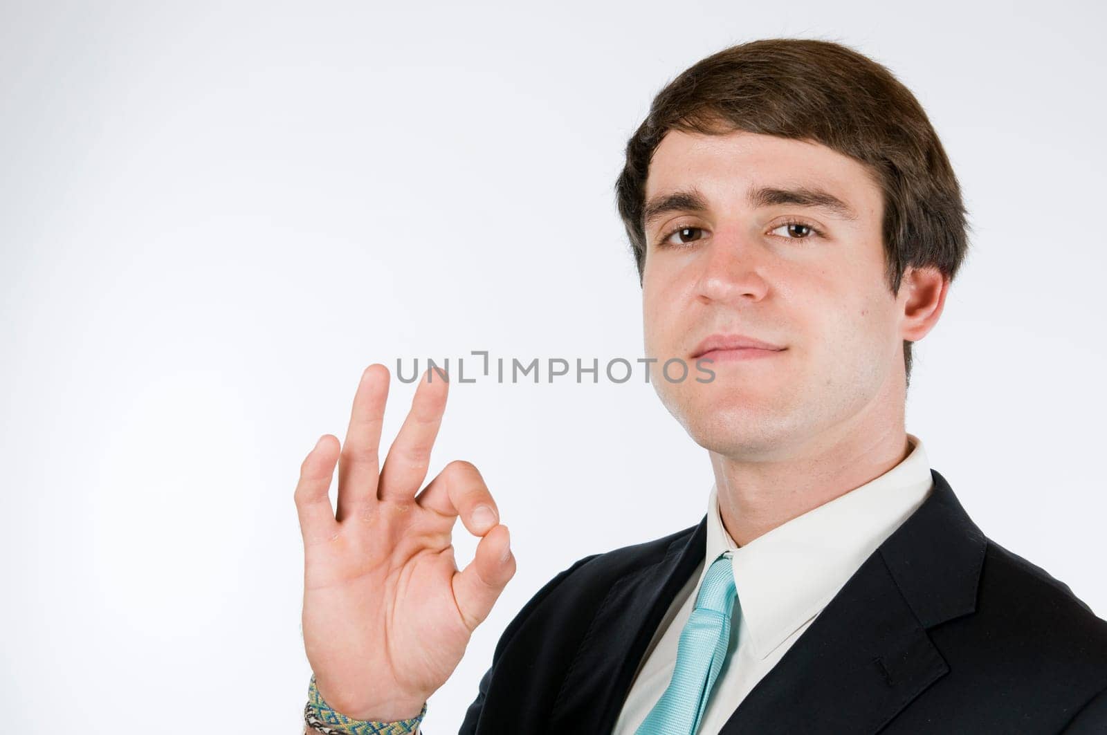 Head-and-shoulder view of a young man around dark suit against a white background showing the O.K. sign and smiling friendly into the camera.
