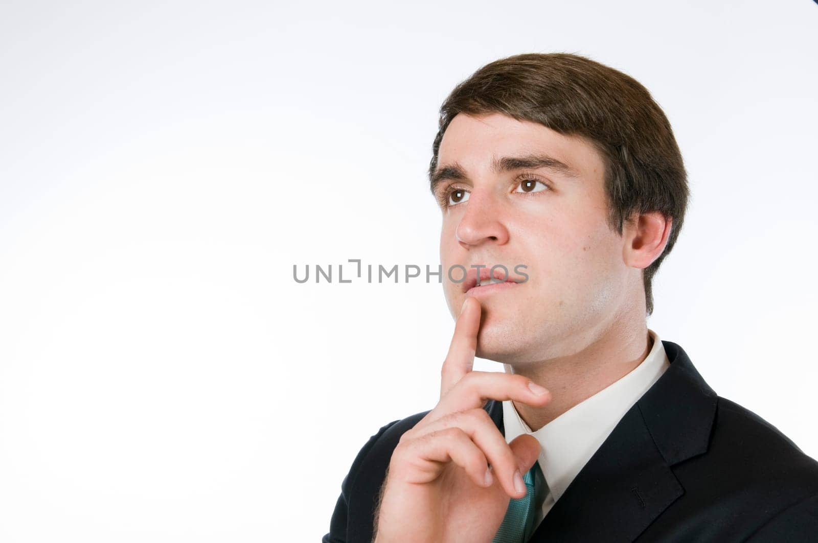 Head-and-shoulder view of a young man around dark suit against a white background,the index finger placed on the chin with a thoughtful look to the top left.