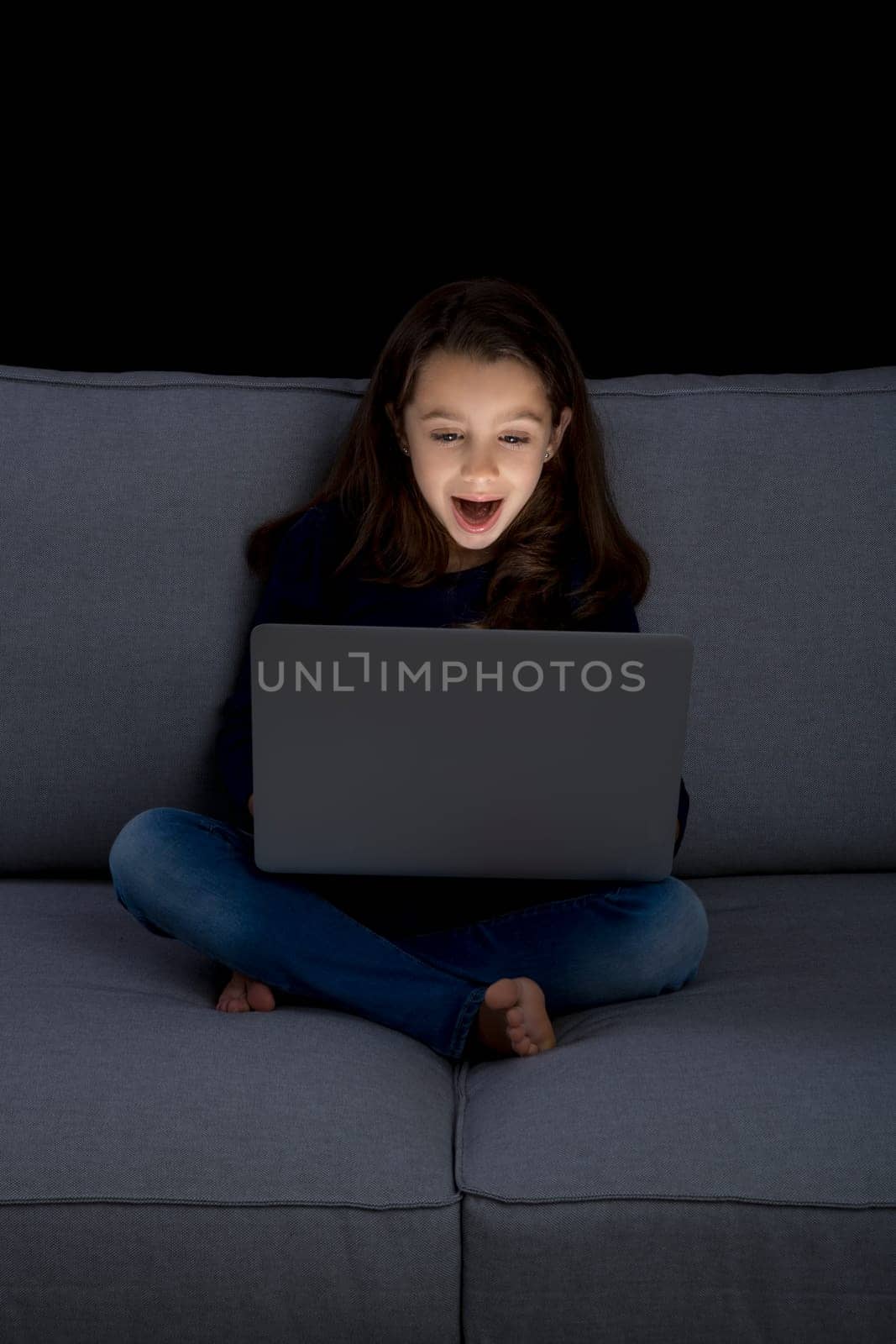 Happy little girl sitting on a couch and working a laptop