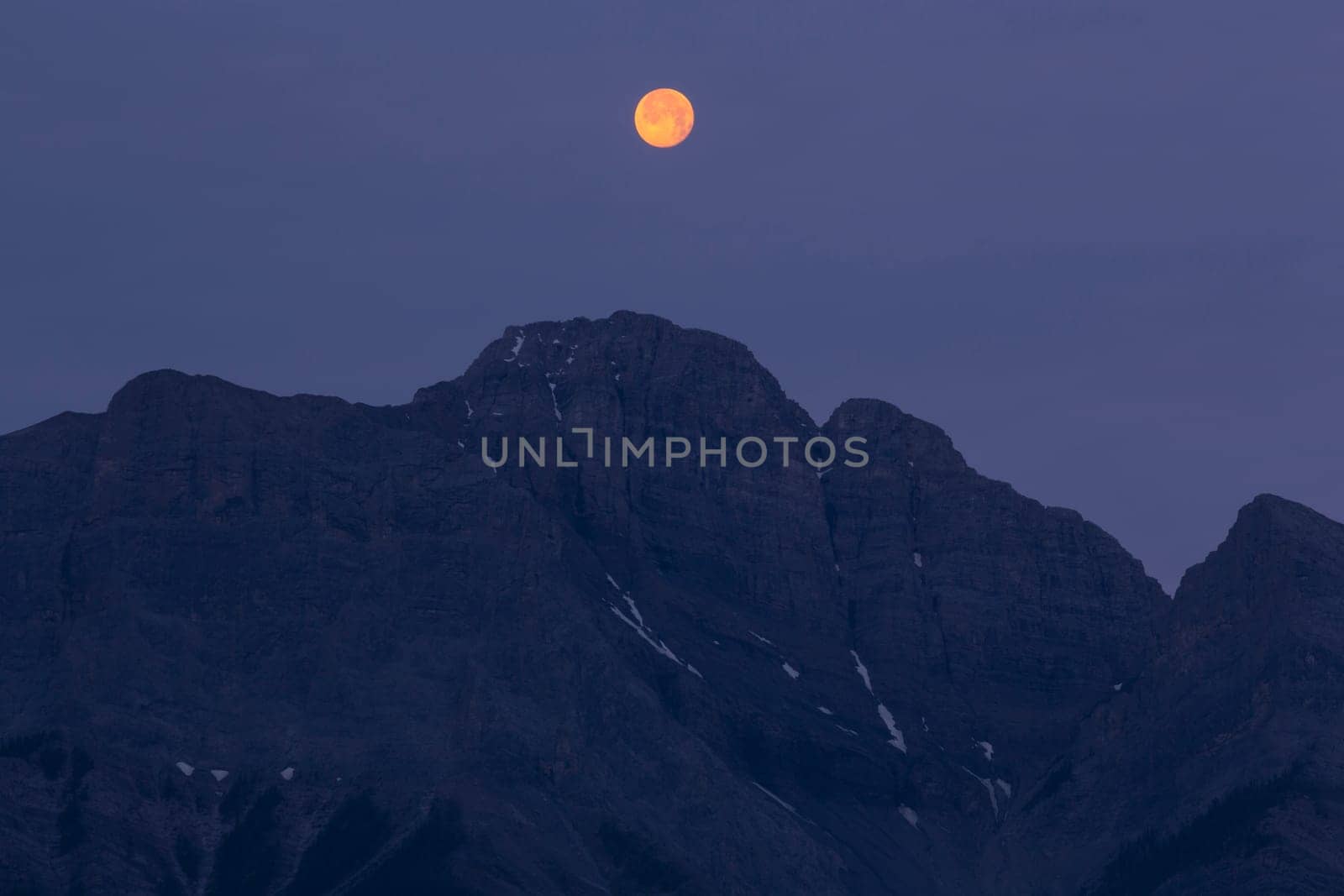 Full moon in early morning sunlight hanging above a mountain range in the Canadian Rocky Mountains, Alberta, Canada