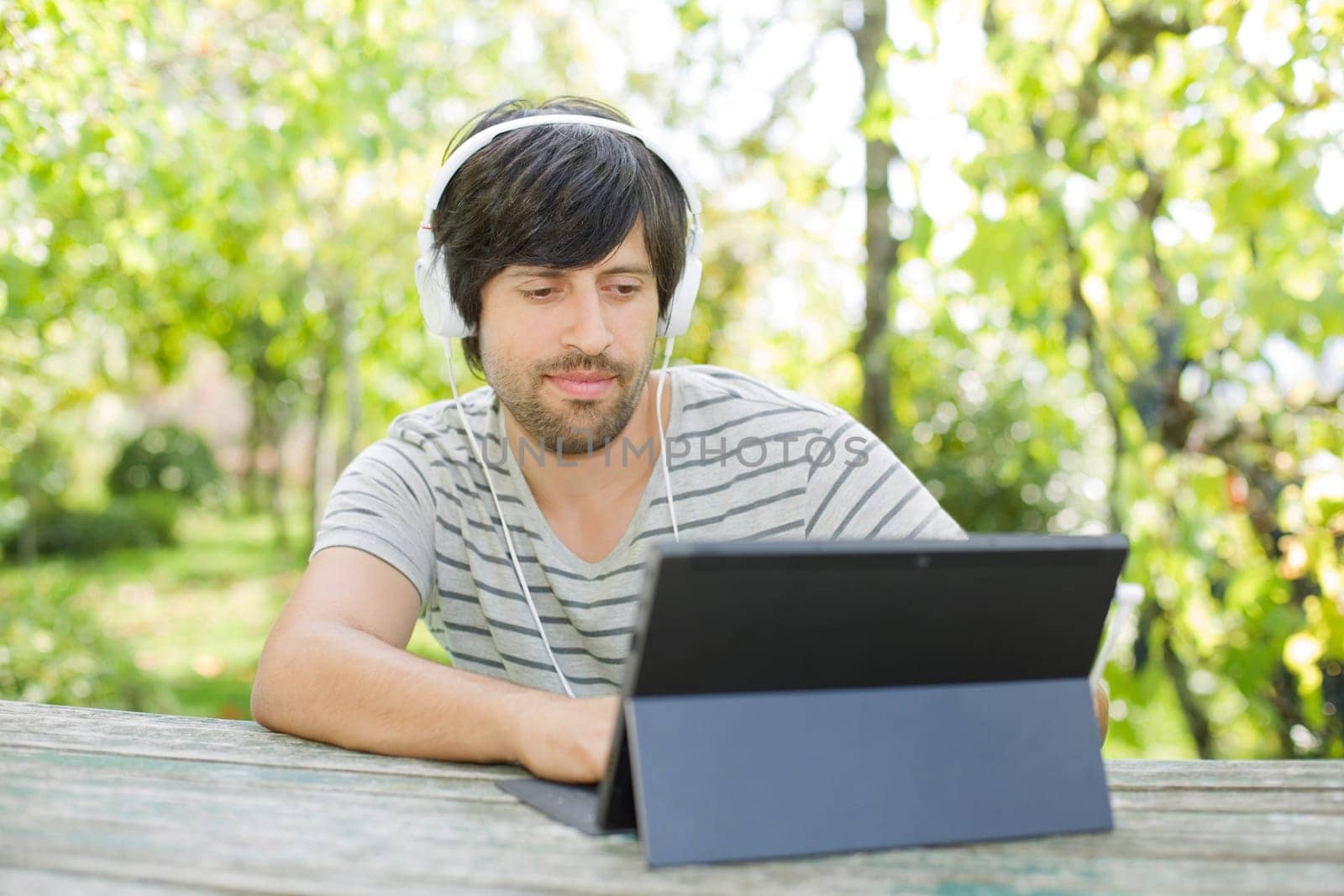 young man working with a tablet pc listening music with headphones on a wooden table, outdoor