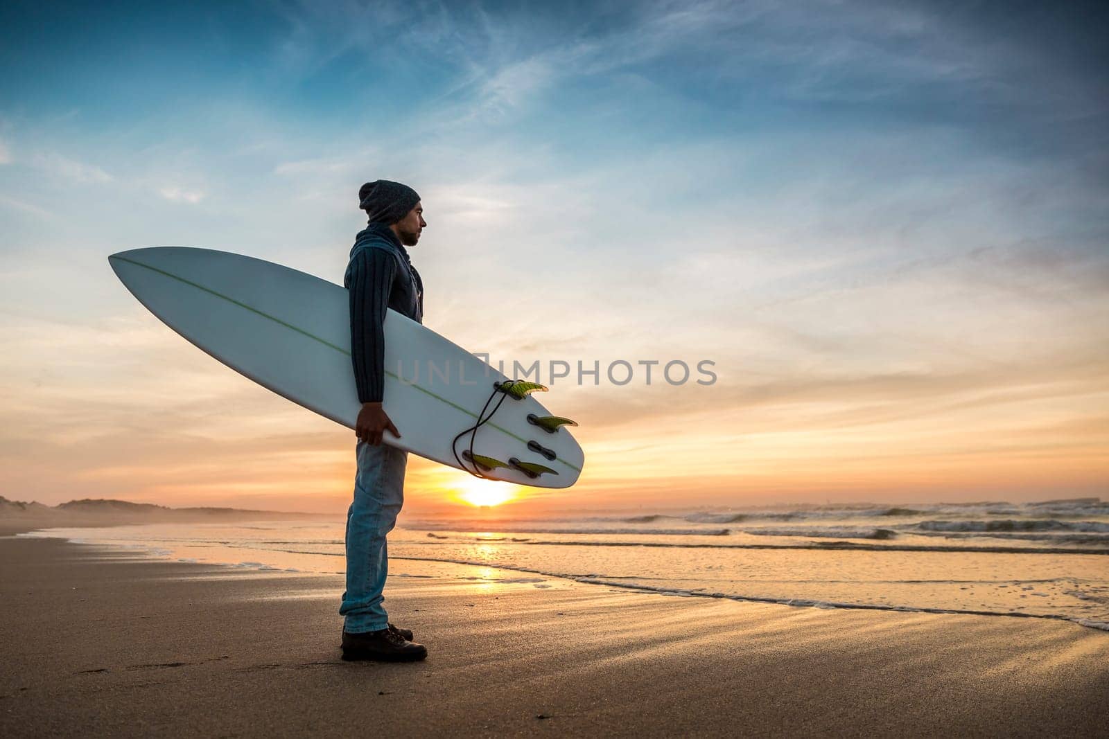A surfer with his surfboard at the sunset looking to the waves