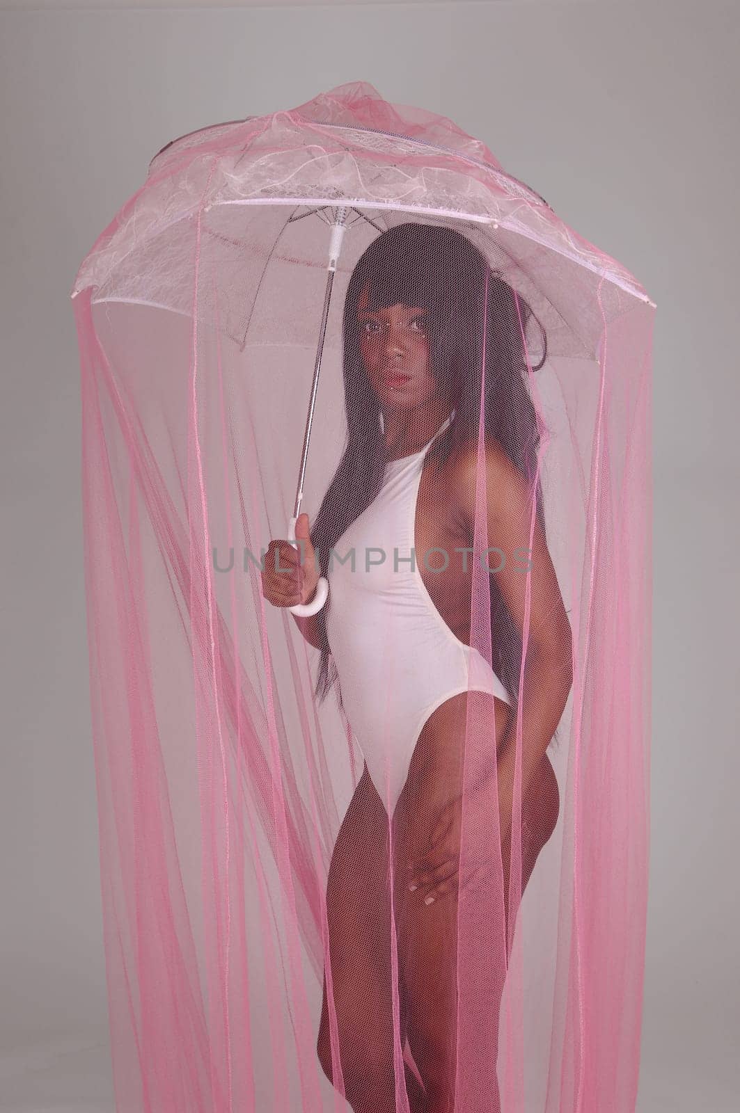A lovely African American woman standing in a white bathing suit under .pink twill, holding an umbrella, isolated for gray background..