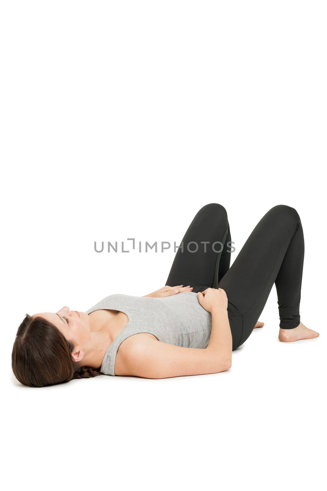 Flat-angled full-body view of a young woman lying on her back in a variant of the yoga position "happy baby"" (anada balasana) on a white background."