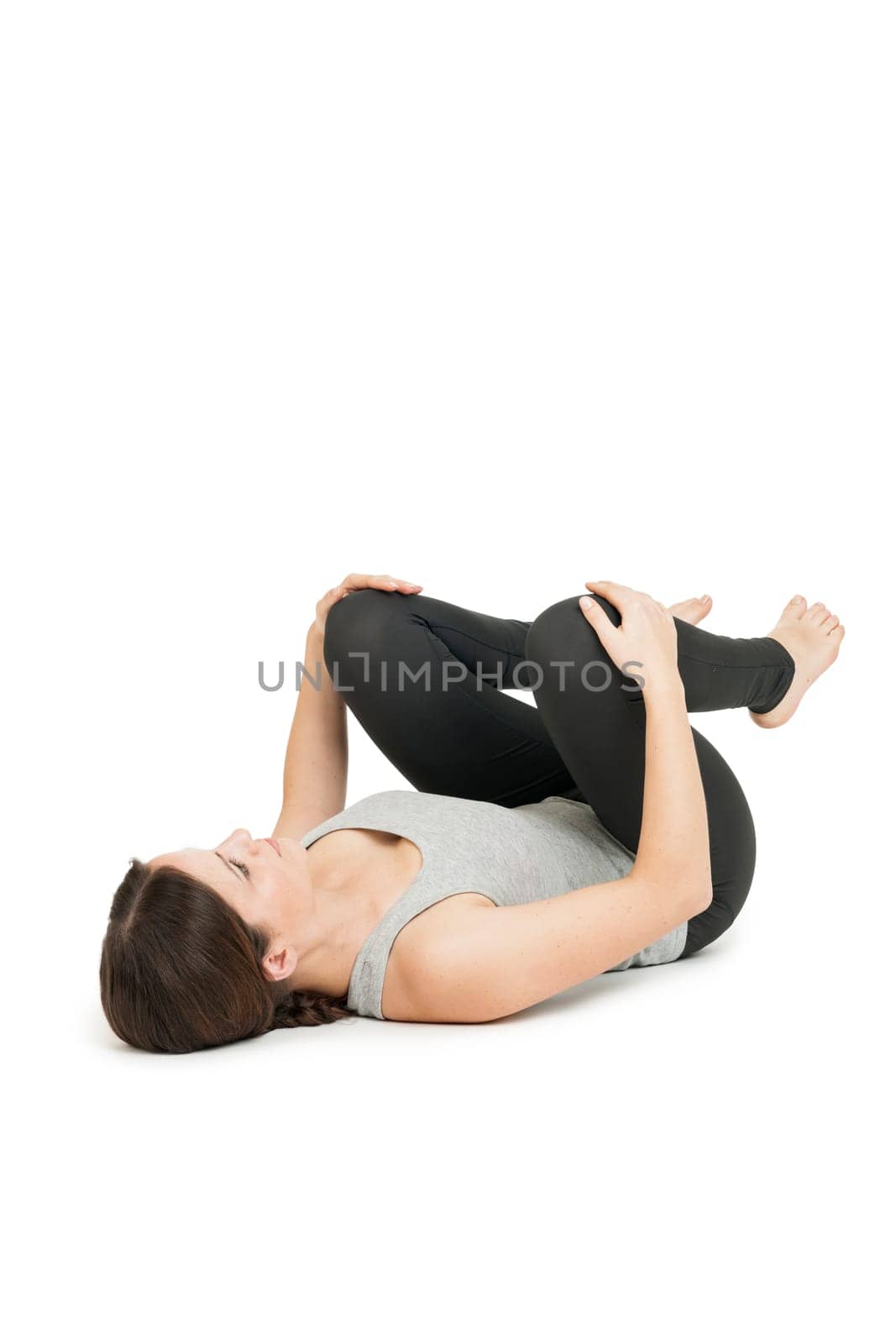 Flat-angled full-body view of a young woman lying on her back in the yoga position "happy baby"" (anada balasana) on a white background."