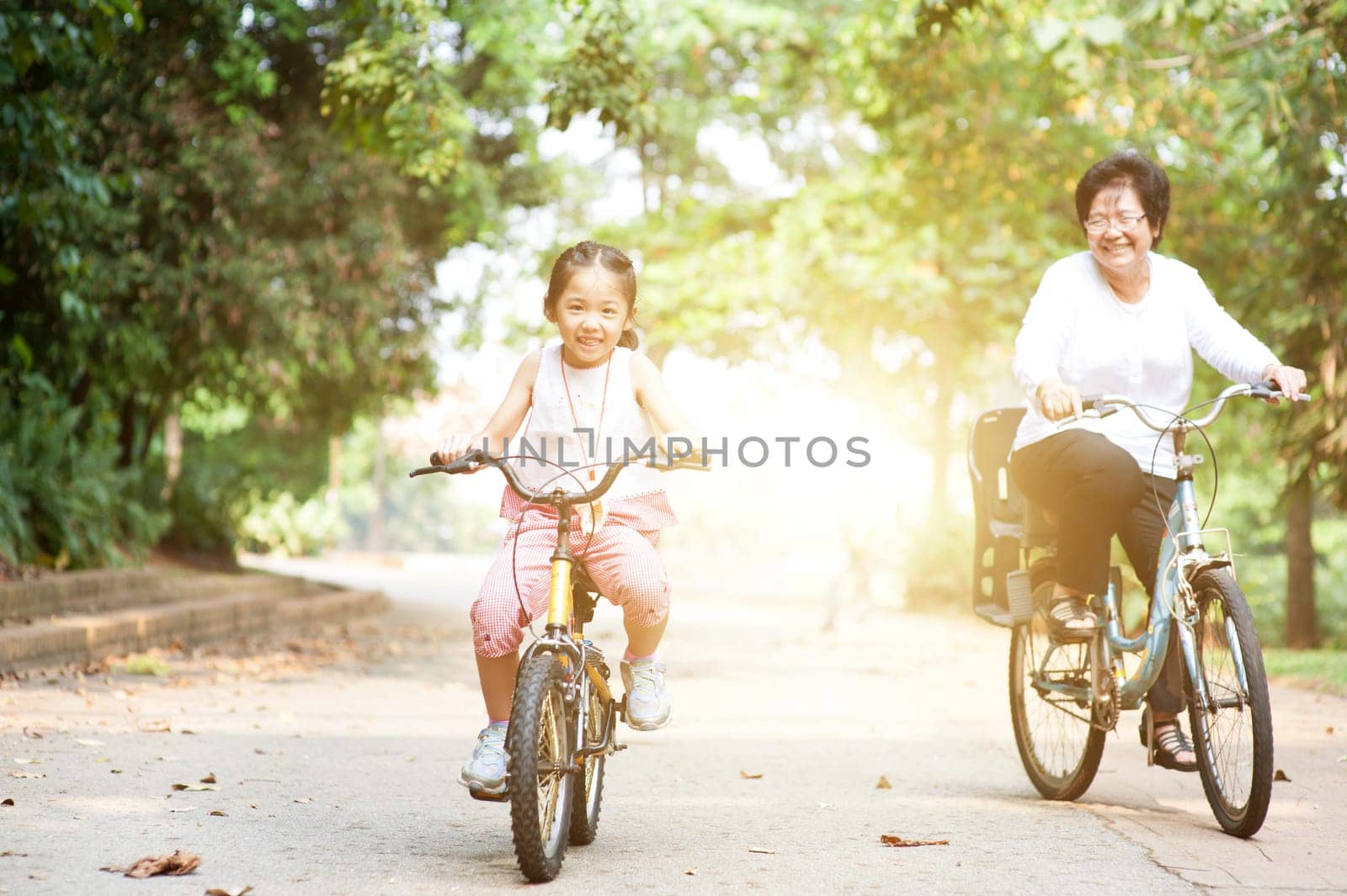 Portrait of active multi generations Asian family at nature park. Grandmother and granddaughter riding bicycle outdoor. Morning sun flare background.