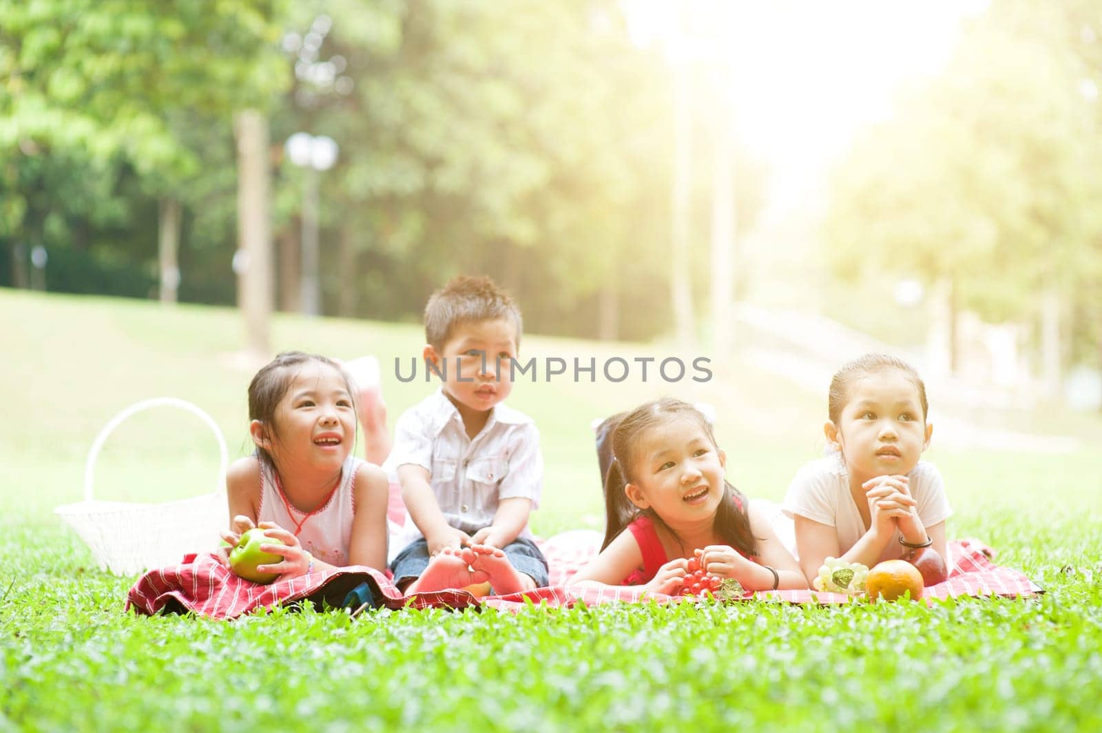 Portrait of Asian children picnics at park, lying on lawn. Little boy and girls having fun outdoors. Morning sun flare background.