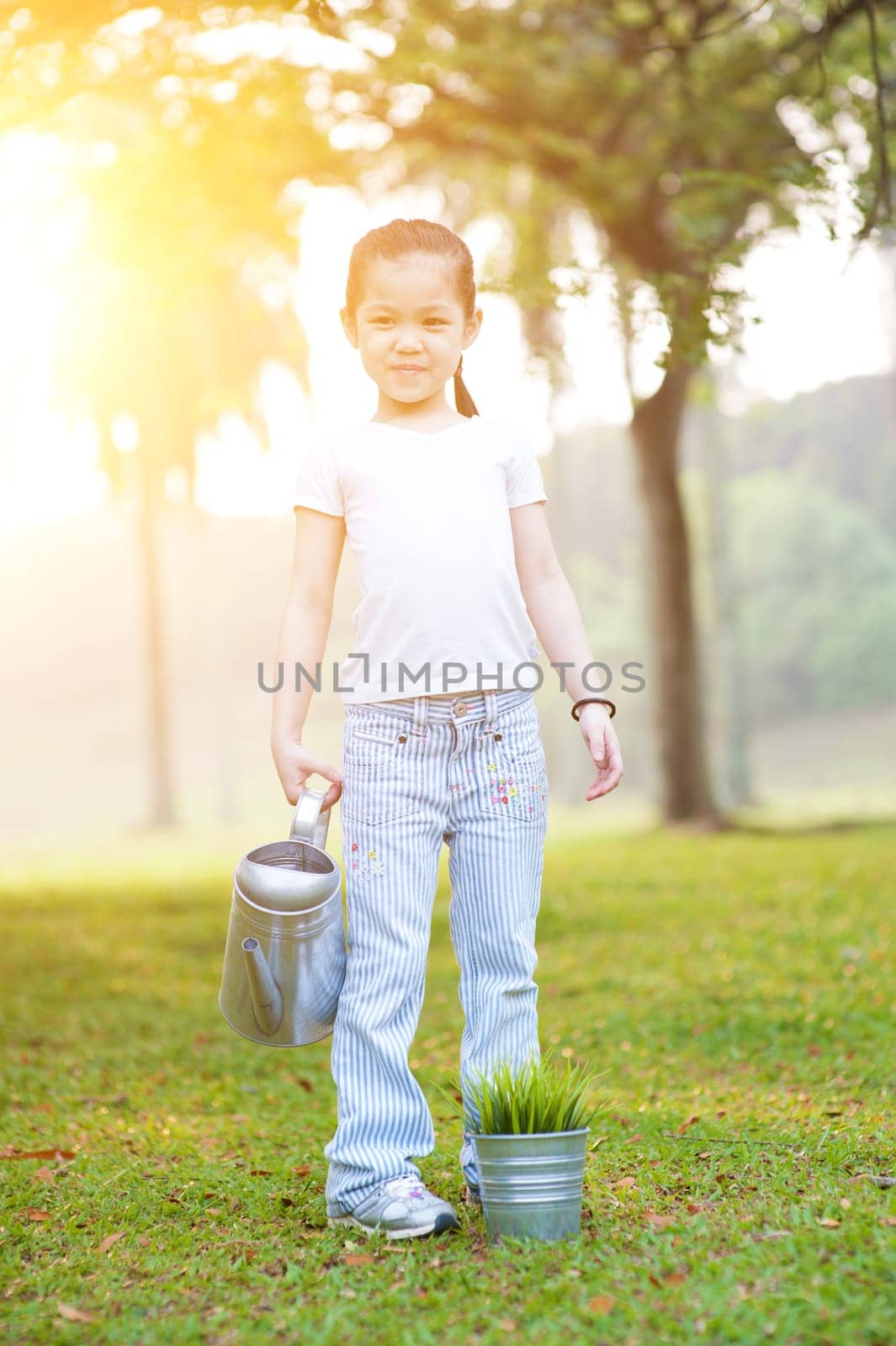 Portrait of cute Asian kid watering plant outdoors. Little girl having fun at nature park. Morning sun flare background.