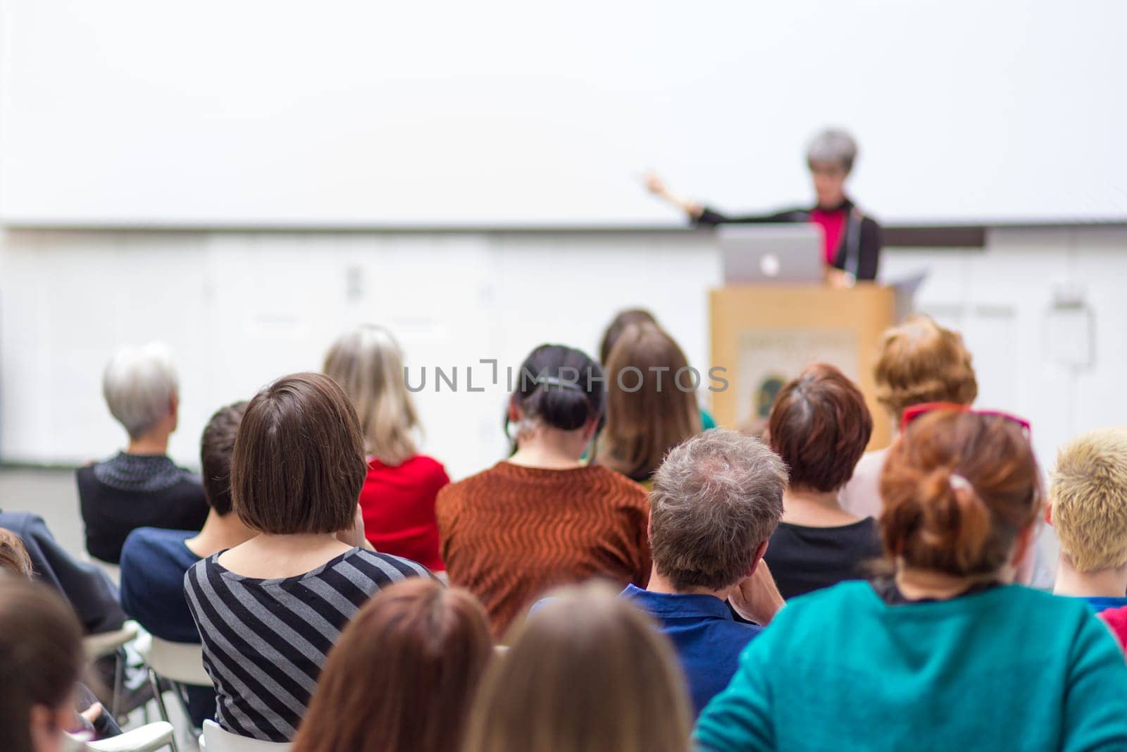 Business and entrepreneurship symposium. Female speaker giving a talk at business meeting. Audience in conference hall. Rear view of unrecognized participant in audience. Copy space on whitescreen.