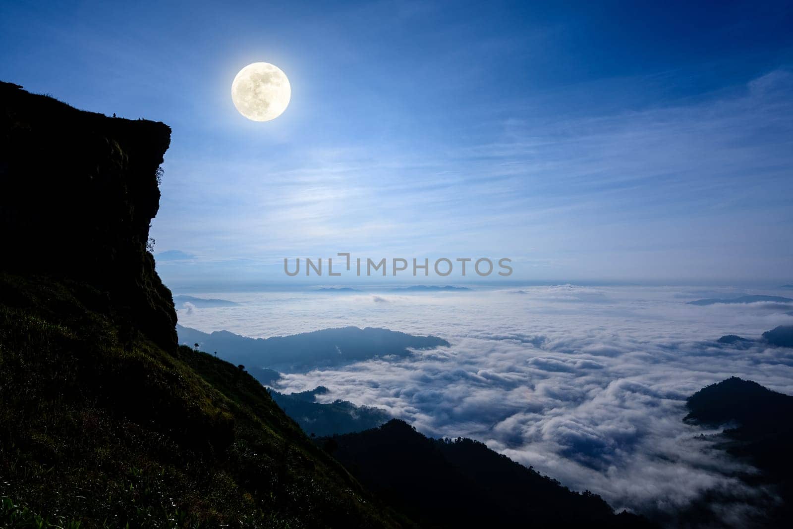 Beautiful landscape nature, Full moon with bright white light over the clouds and fog in the dark blue sky at night on the peak mountain in winter at Phu Chi Fa Forest Park, Chiang Rai Province, Thailand