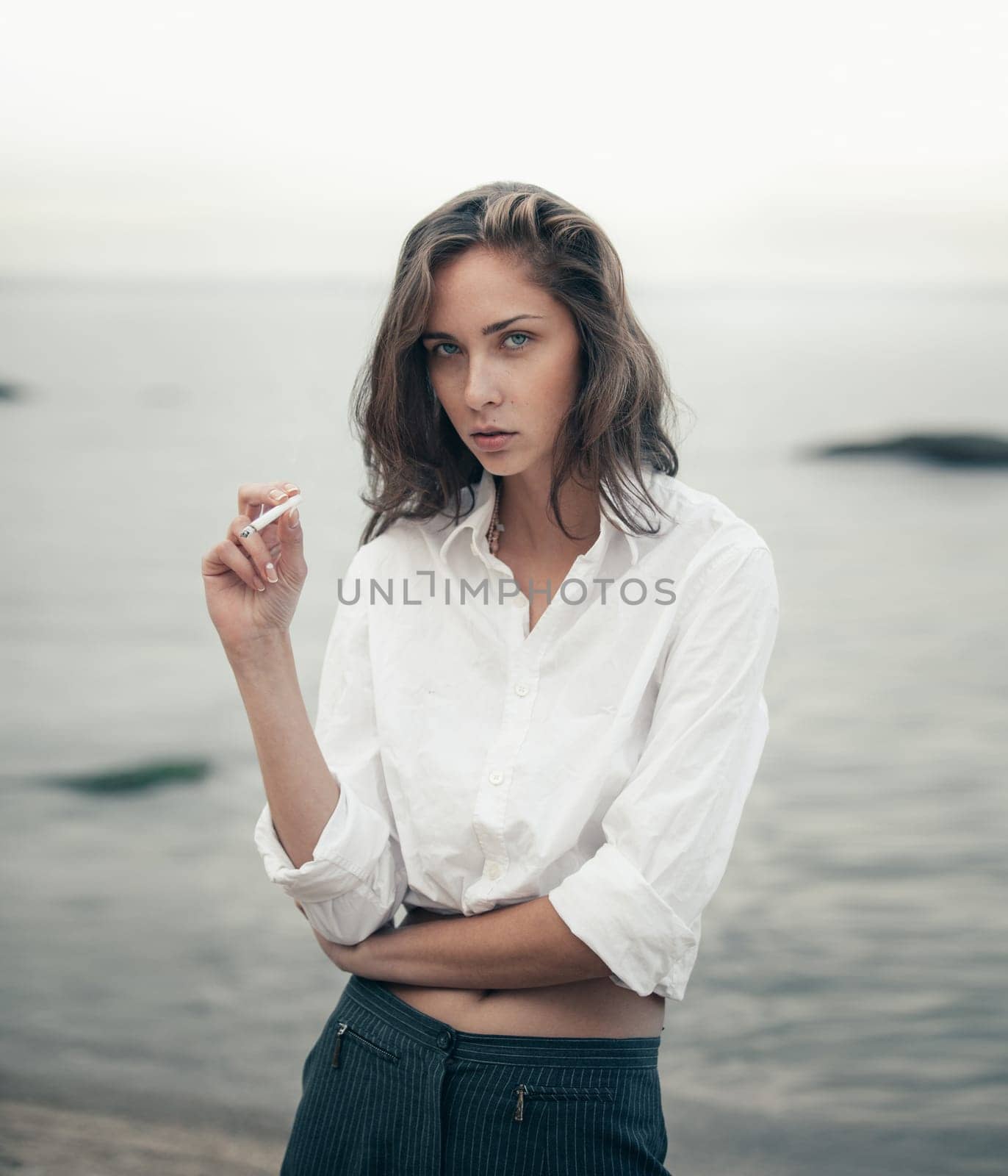 Beautiful business woman in white shirt and pants smokes a cigarette on the beach near the sea.