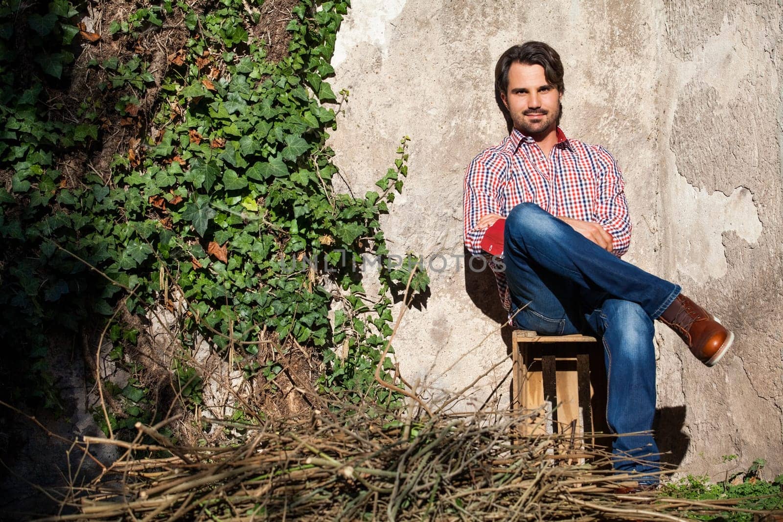 Young man with a plaid shirt and jeans with dark hair and beard outdoors under an efeuranke in daylight