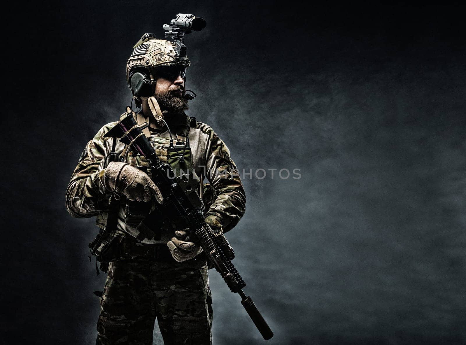Bearded soldier in Combat Uniforms with weapon, plate carrier and combat helmet are on. Studio shot, dark background
