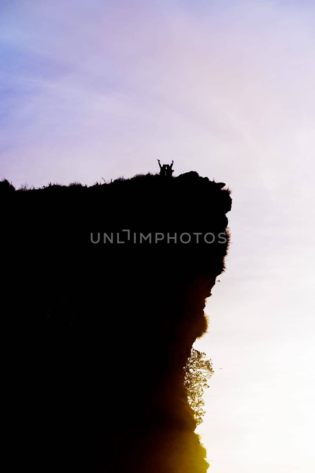 Black silhouette on sky background of two tourist standing arms outstretched happy with success on peak mountain near the cliff during sunrise at Phu Chi Fa Viewpoint, Chiang Rai, Thailand