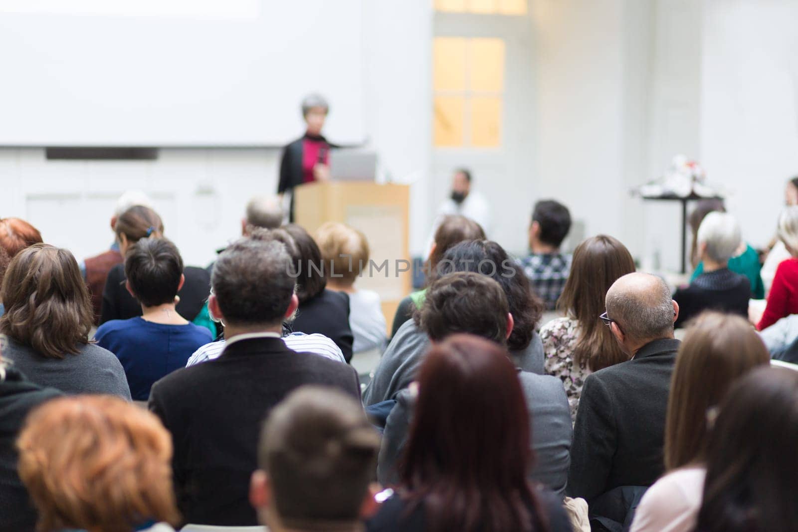 Business and entrepreneurship symposium. Female speaker giving a talk at business meeting. Audience in conference hall. Rear view of unrecognized participant in audience.
