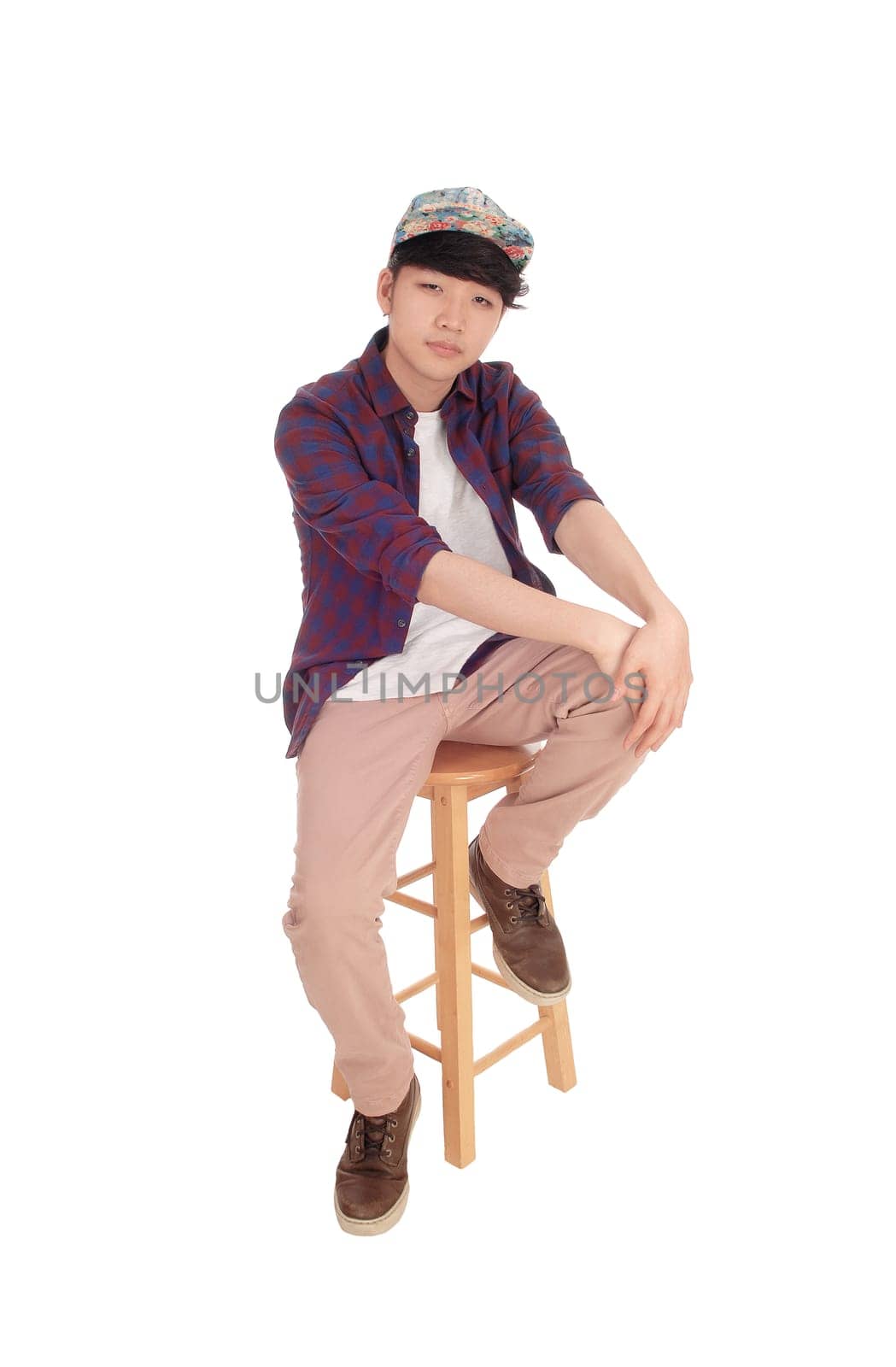 A young Korean teenager sitting on a chair with a cap with his.hands on his knee, isolated for white background..