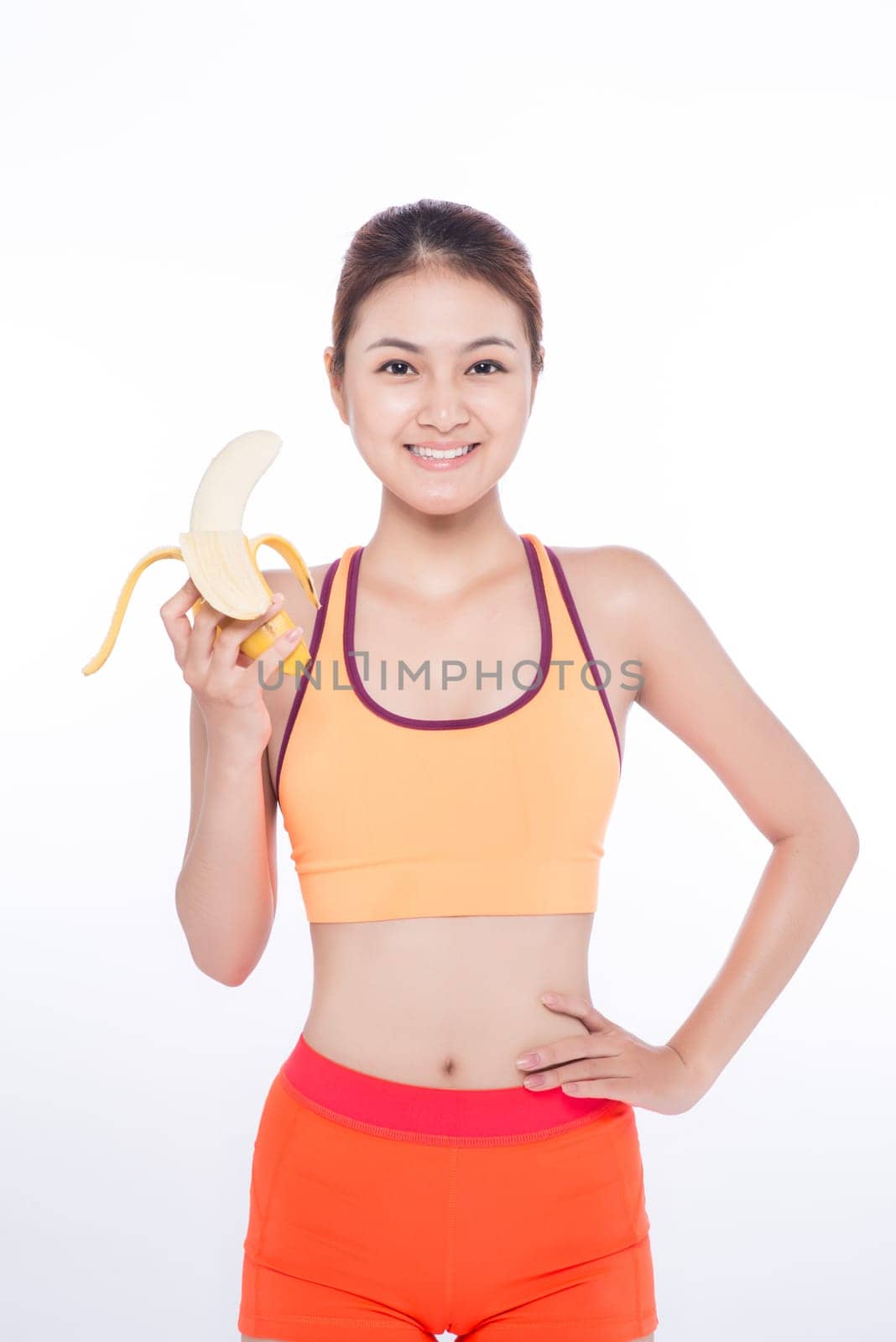 Smiling sporty asian  woman holding banana over white background and looking at camera
