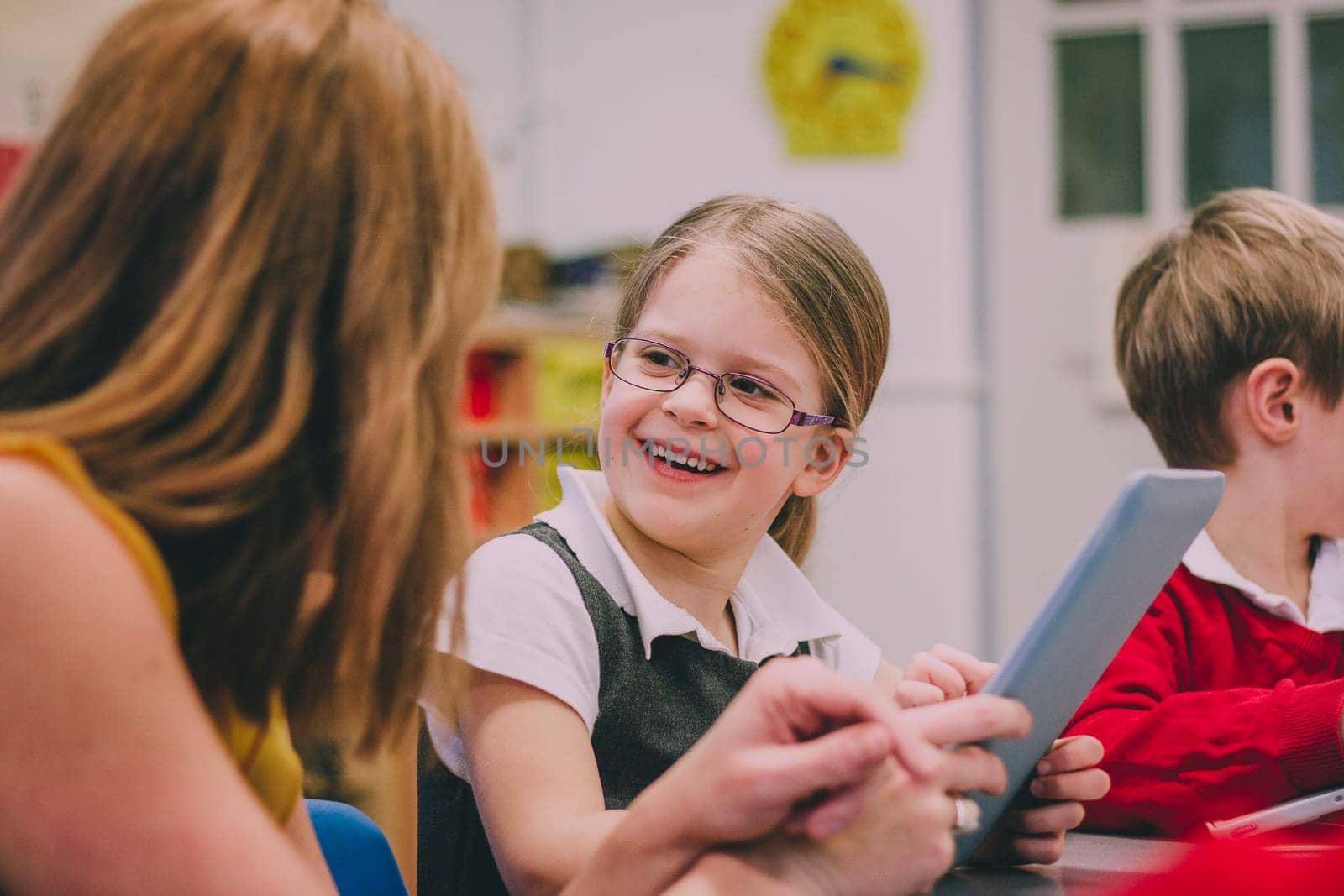 Primary school students are using digital tablets in their classroom lesson. A little girl is looking at her teacher and laughing.